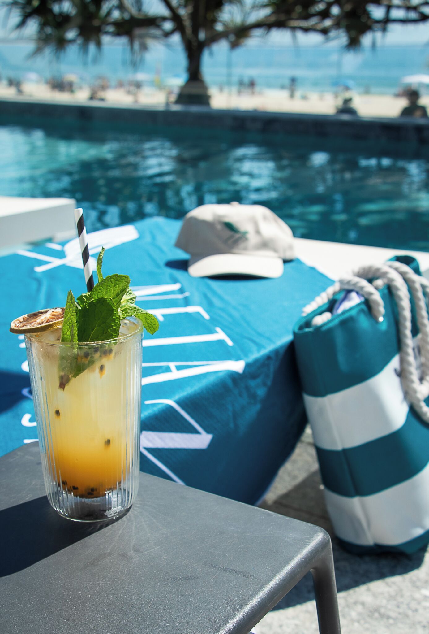 On aperçoit clairement une zone autour d’une piscine, avec un verre contenant une boisson jaune, un sac à rayures et une chaise longue bleue et blanche.