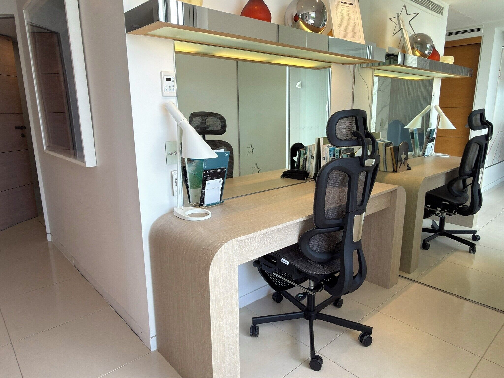 A modern office space with a desk, two office chairs, a lamp, a mirror, and a bookshelf.