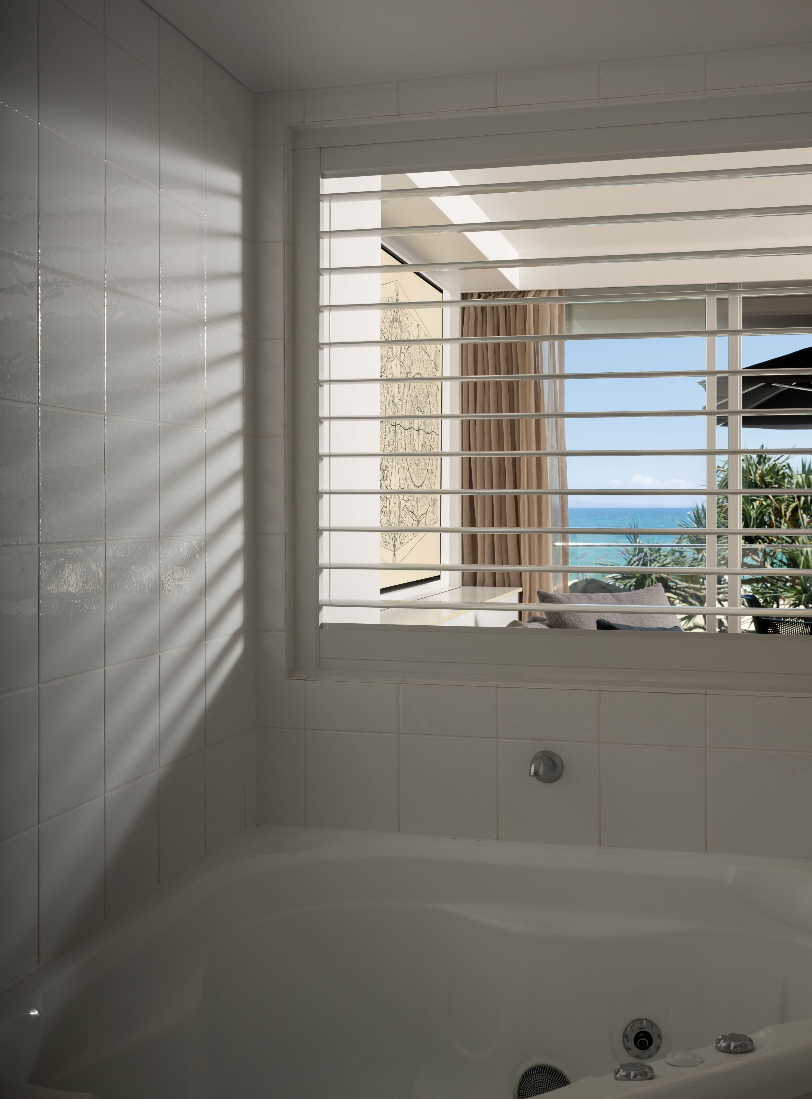 A modern bathroom with a large bathtub, a window with blinds, and a view of the ocean.