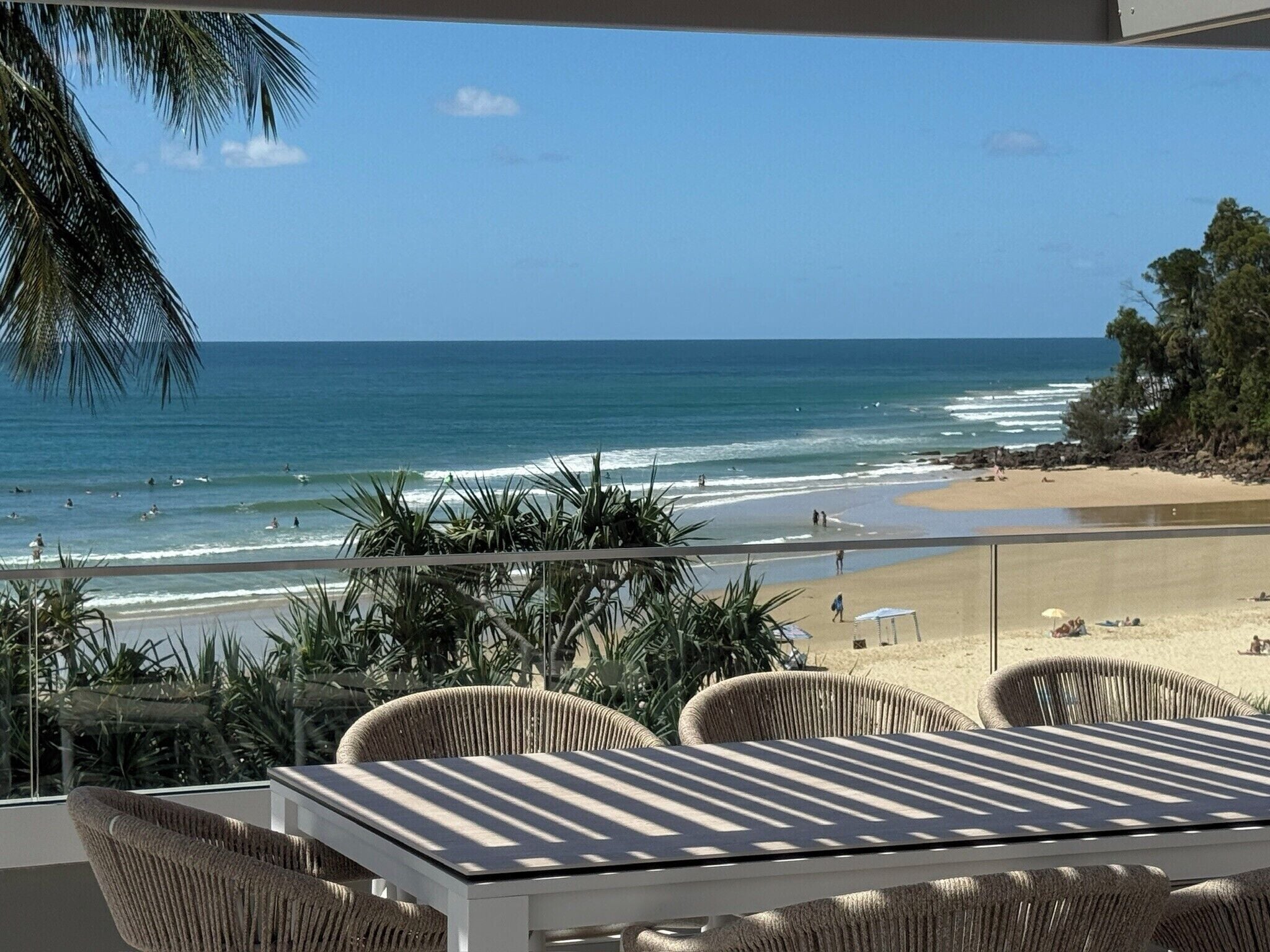 A balcony with wicker furniture overlooking a beach with waves and a clear sky.