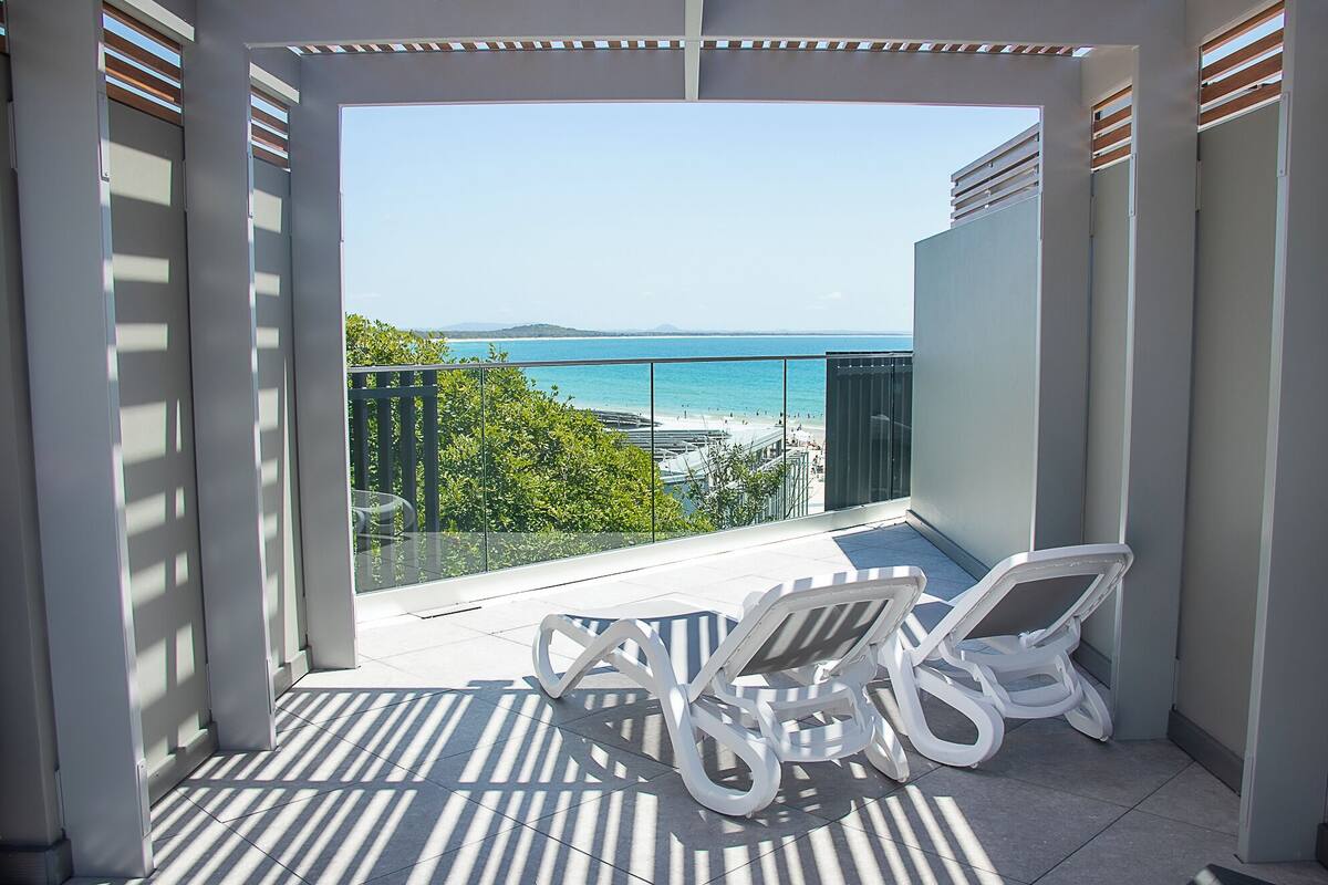 A balcony with two white lounge chairs, a glass balustrade, and a view of a beach and sea.