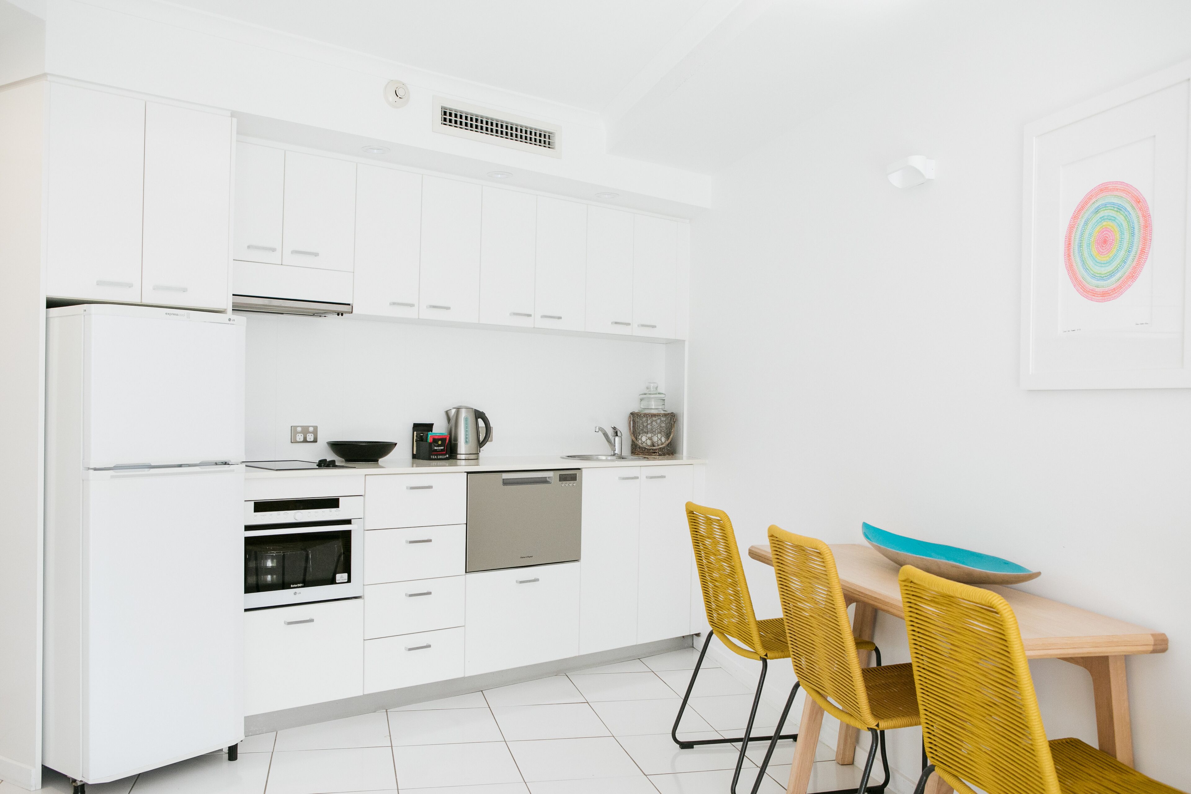 Una cocina moderna con armarios blancos, lavavajillas, horno y comedor con mesa de madera y sillas amarillas.