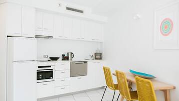 Una cocina moderna con armarios blancos, lavavajillas, horno y comedor con mesa de madera y sillas amarillas.