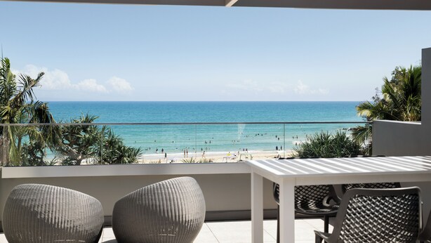 Een balkon met rieten stoelen en een tafel met uitzicht op een strand met mensen en palmbomen.