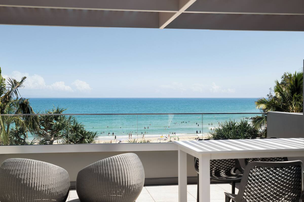 A balcony with wicker chairs and a table overlooking a beach with people and palm trees.