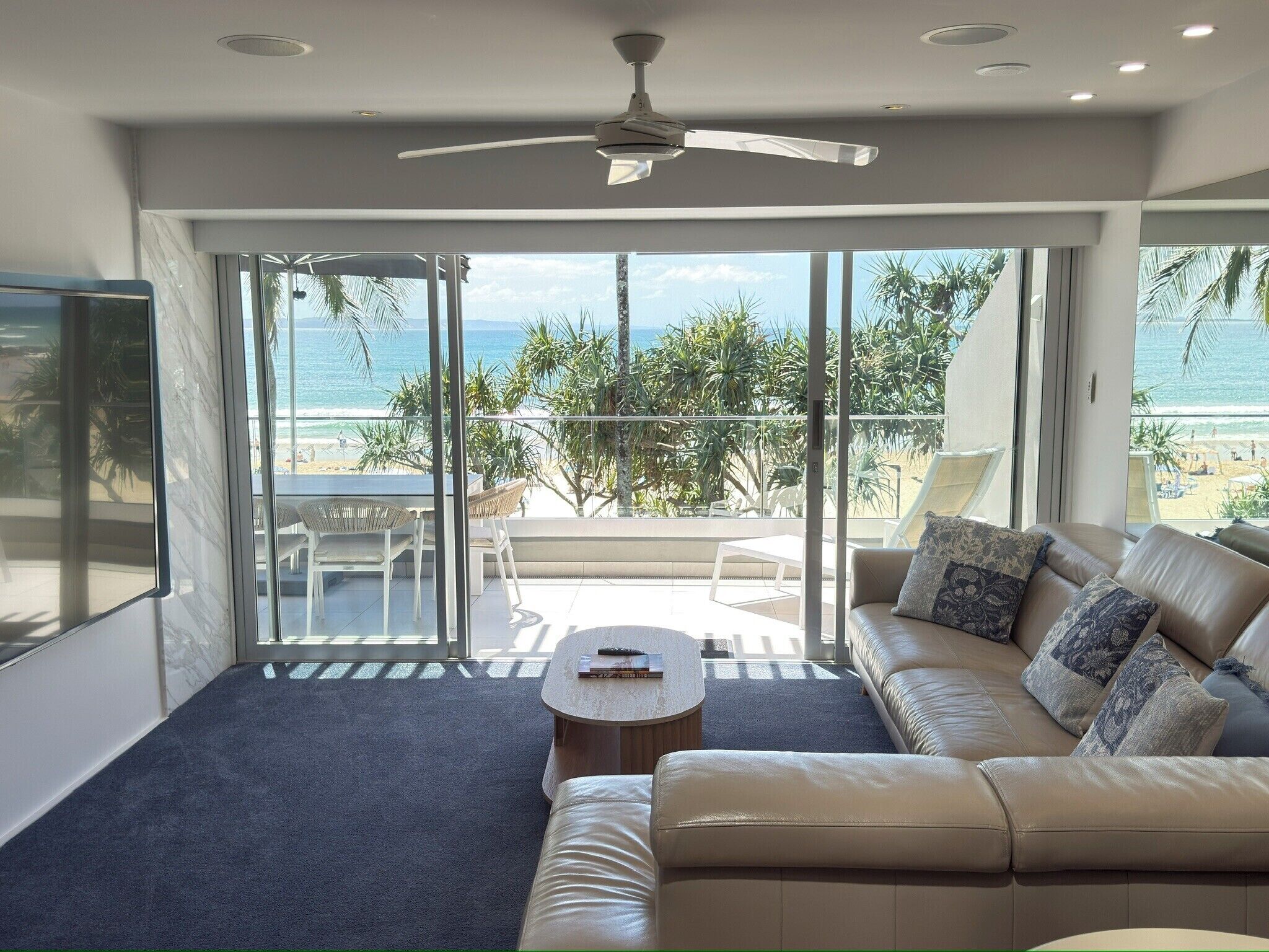A modern living room with a sofa, coffee table, and a ceiling fan. Large glass doors lead to a balcony with a view of the beach.