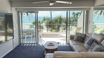 A modern living room with a sofa, coffee table, and a ceiling fan. Large glass doors lead to a balcony with a view of the beach.