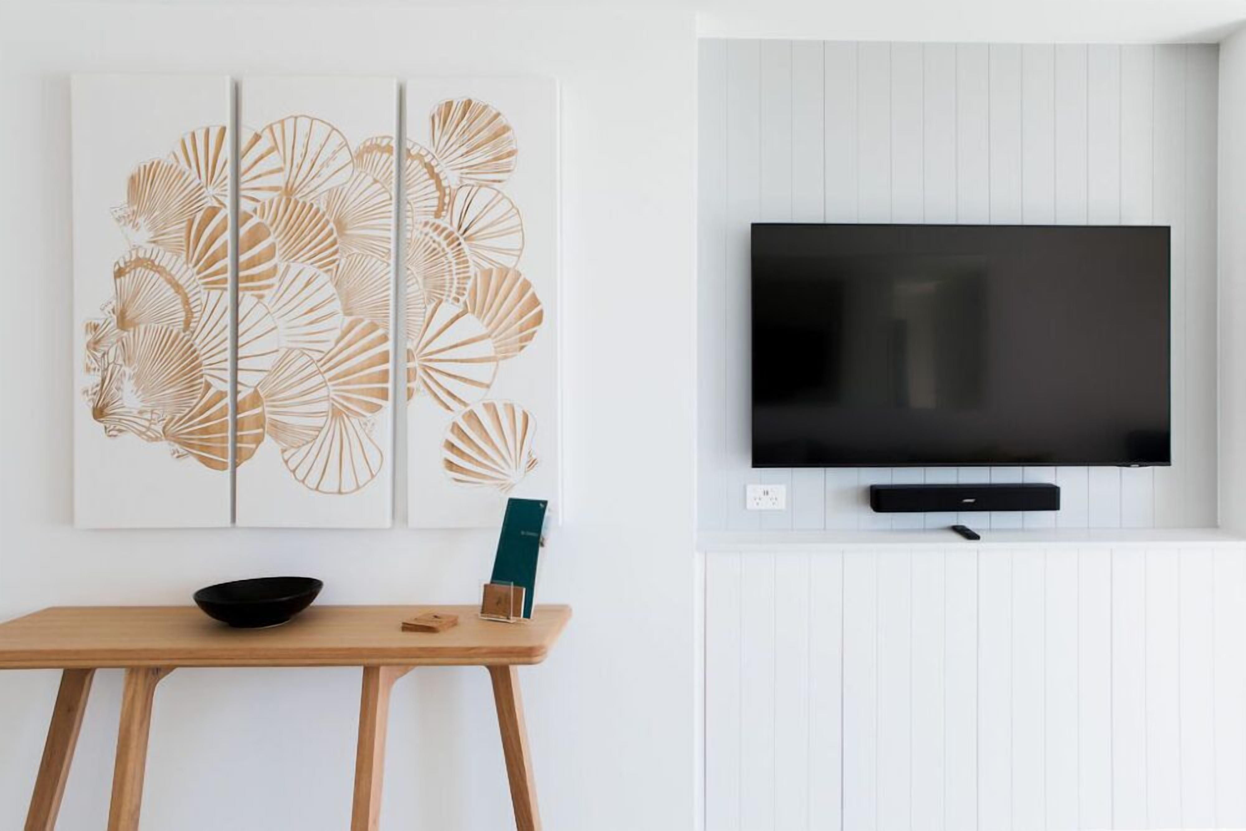 A modern living room with a flat-screen TV mounted on a white wall, a wooden table with a black bowl, and a decorative wall art piece.