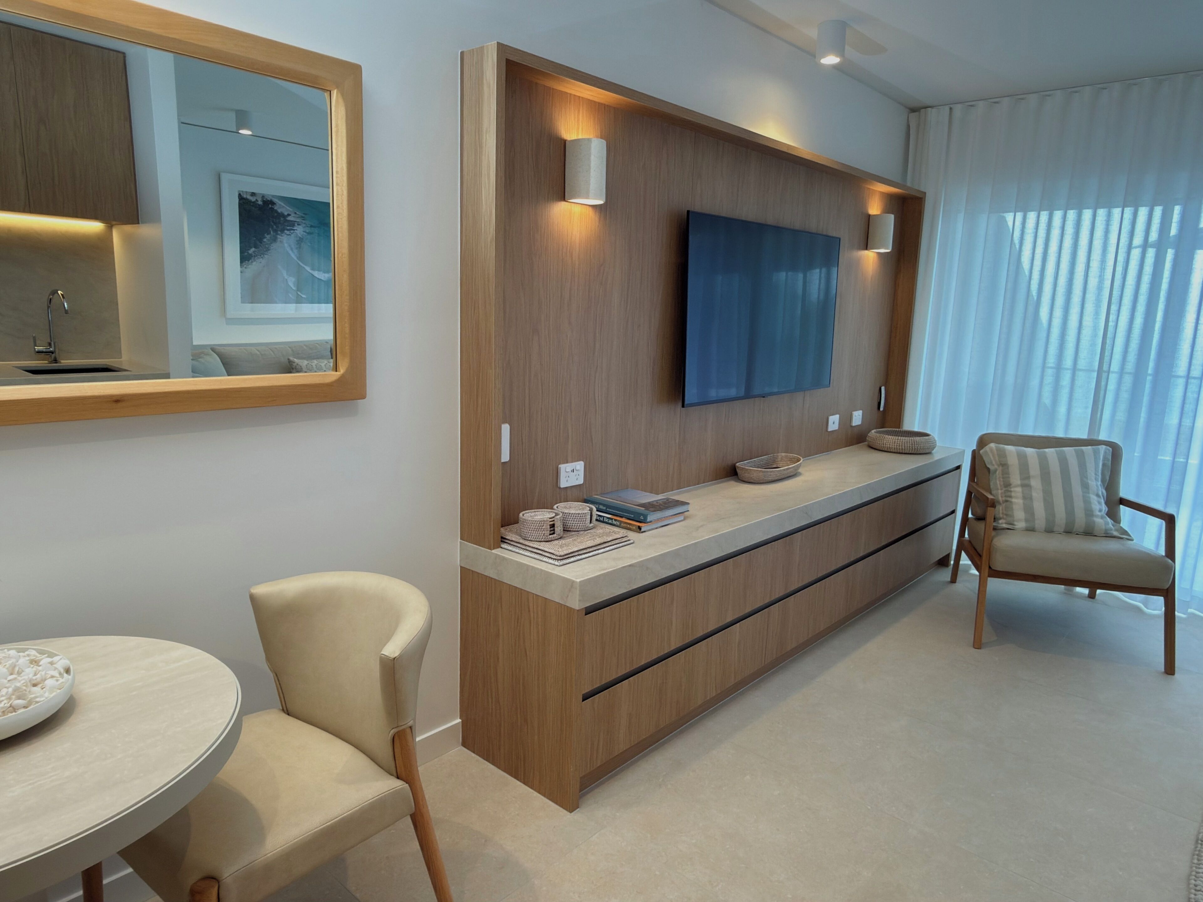 A modern hotel room with a wooden console table, a flat-screen TV, a large mirror, a round table with a bowl of snacks, and a cushioned armchair.