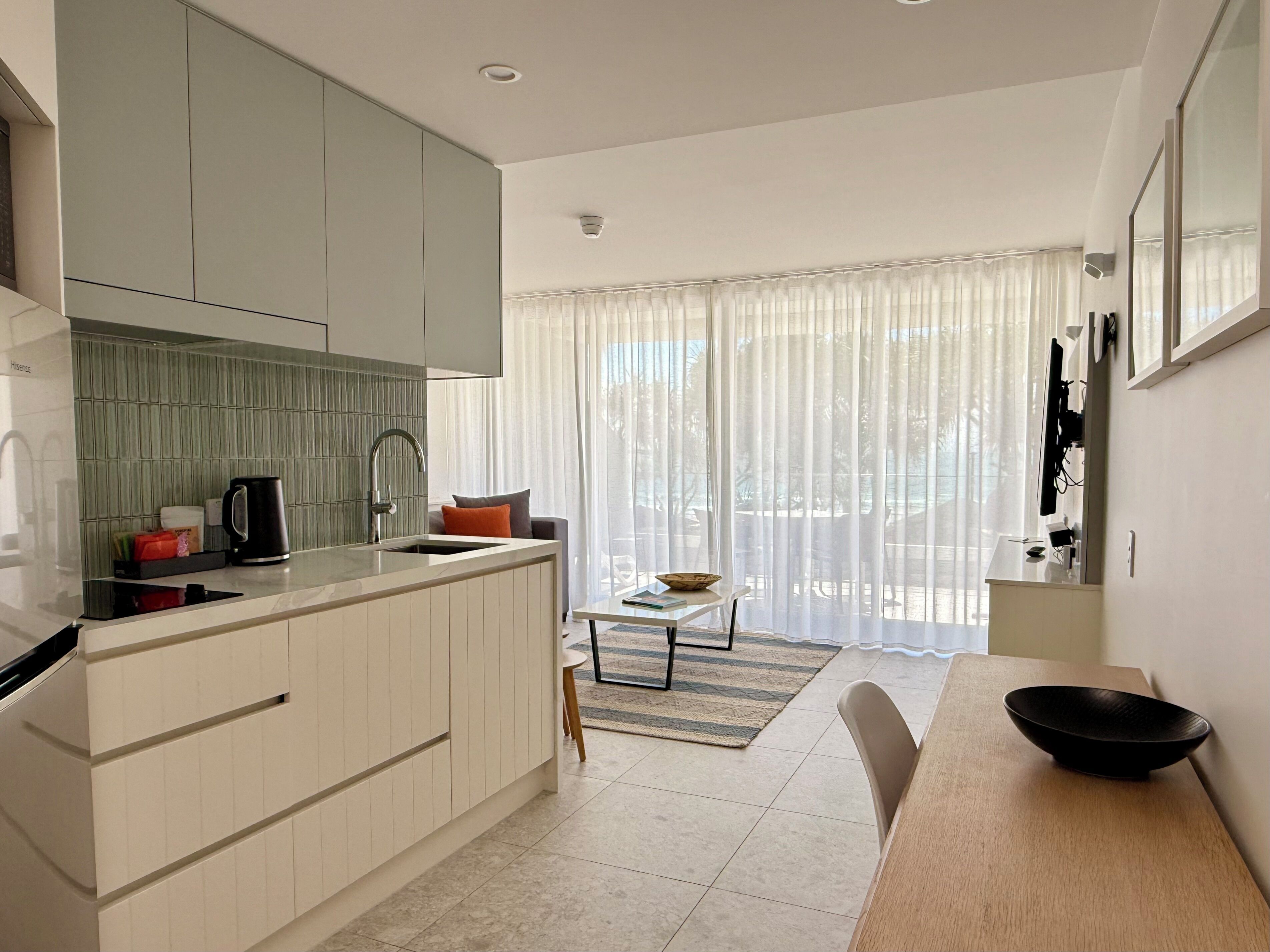 Eine moderne Küche mit integrierter Kücheninsel, einem Essbereich mit Tisch und Stühlen sowie einem großen Fenster mit leichtem Vorhang.