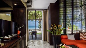 Una sala de estar moderna con un sofá, una mesa de centro y un televisor. Se observa una ventana grande con vista a la playa y palmeras.