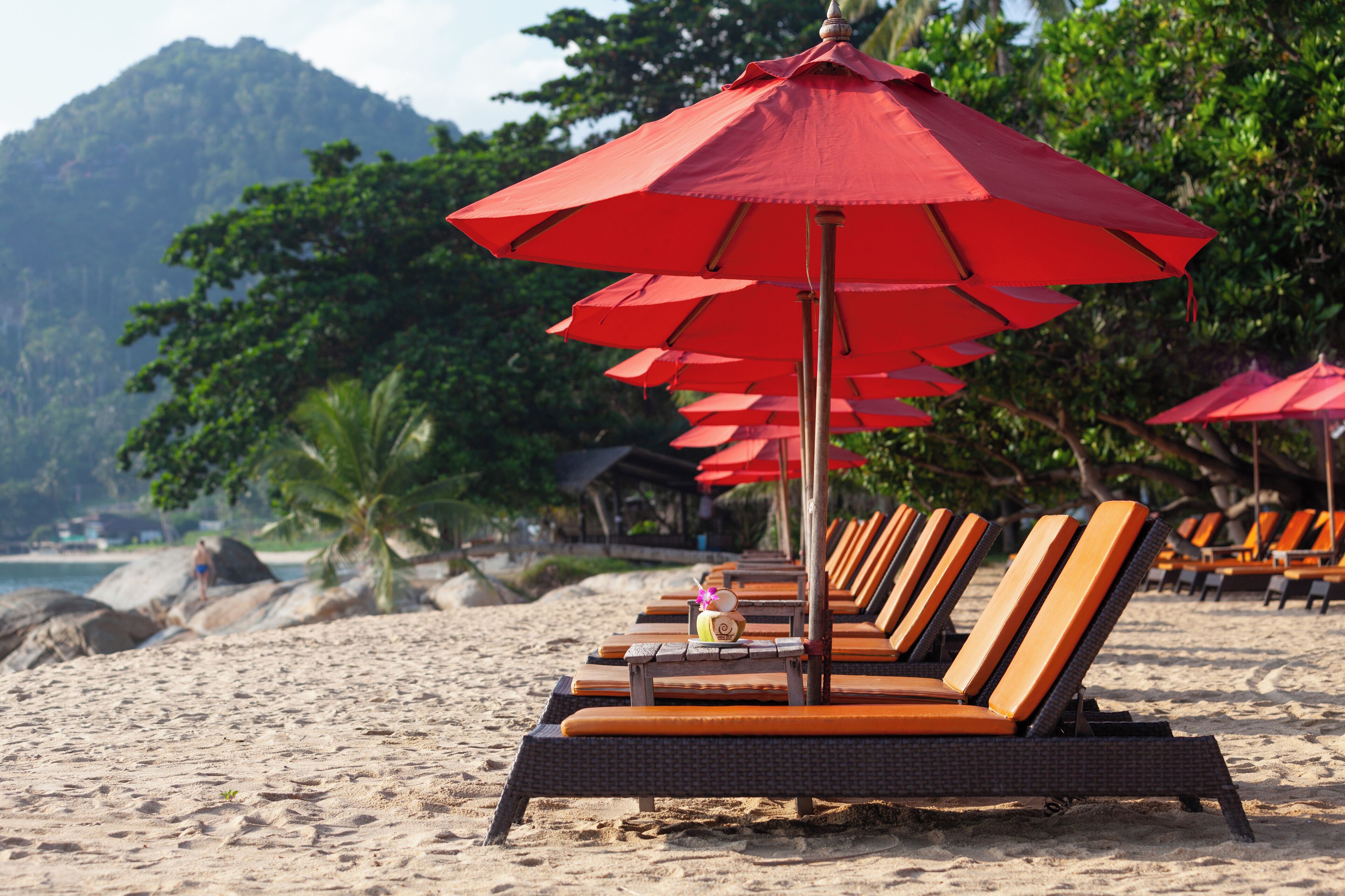 A beach with rows of lounge chairs and red umbrellas, a sandy shore, and a mountain in the background.