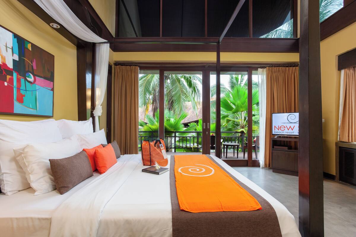 A hotel room with a bed, pillows, a blanket, a TV, and a view of palm trees and a beach.