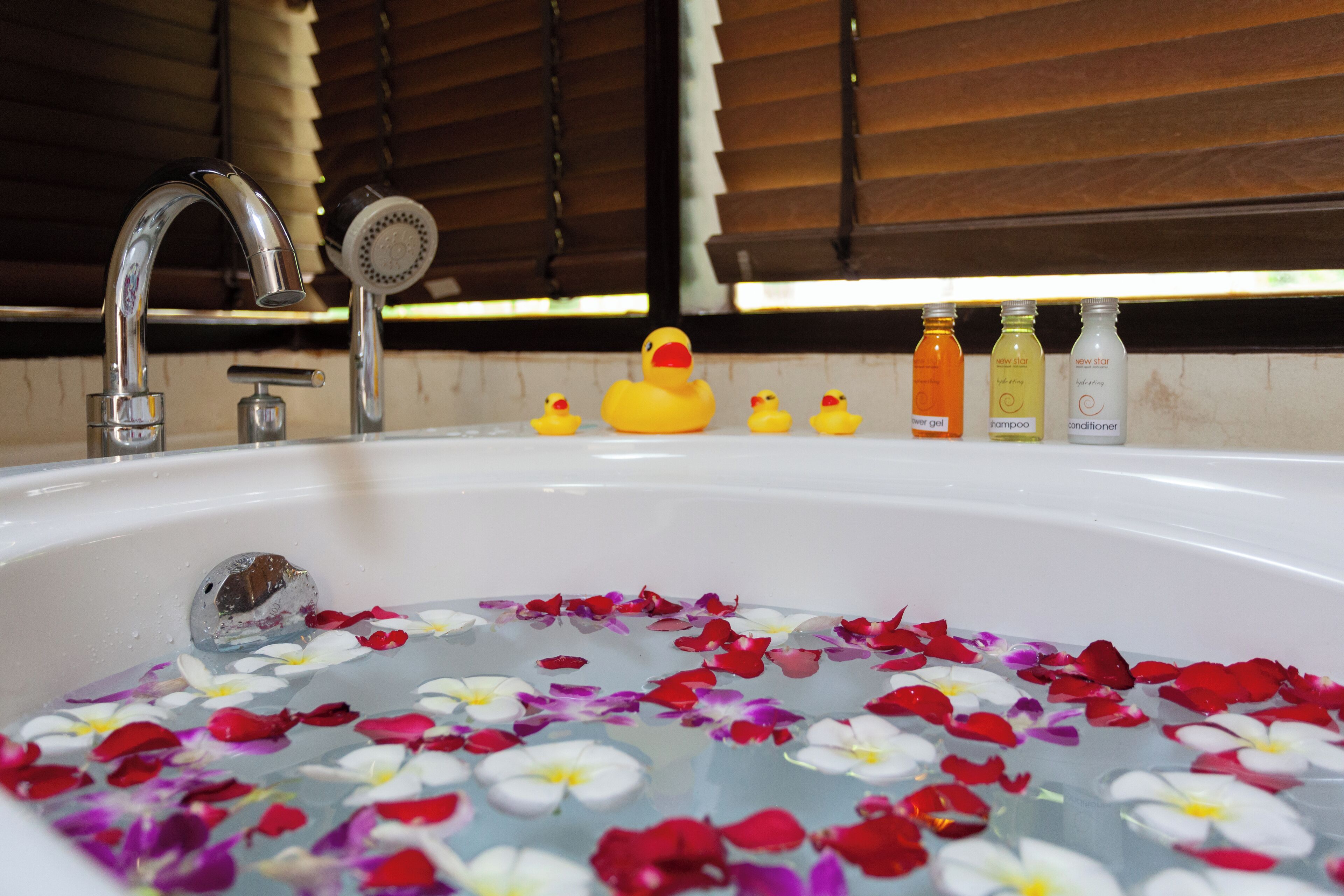 Eine mit Wasser und Rosenblättern gefüllte Badewanne, mit einem quietschenden Badeenten und Flaschen mit Shampoo und Conditioner am Rand.