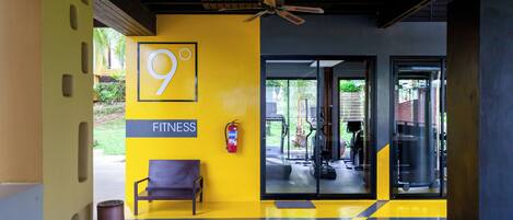 A fitness center entrance with a yellow wall, a bench, a ceiling fan, and a glass door leading to the gym area.