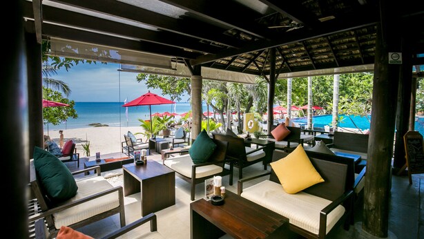 Un espacio al aire libre con mobiliario de madera, cojines y vistas a la playa y al océano.