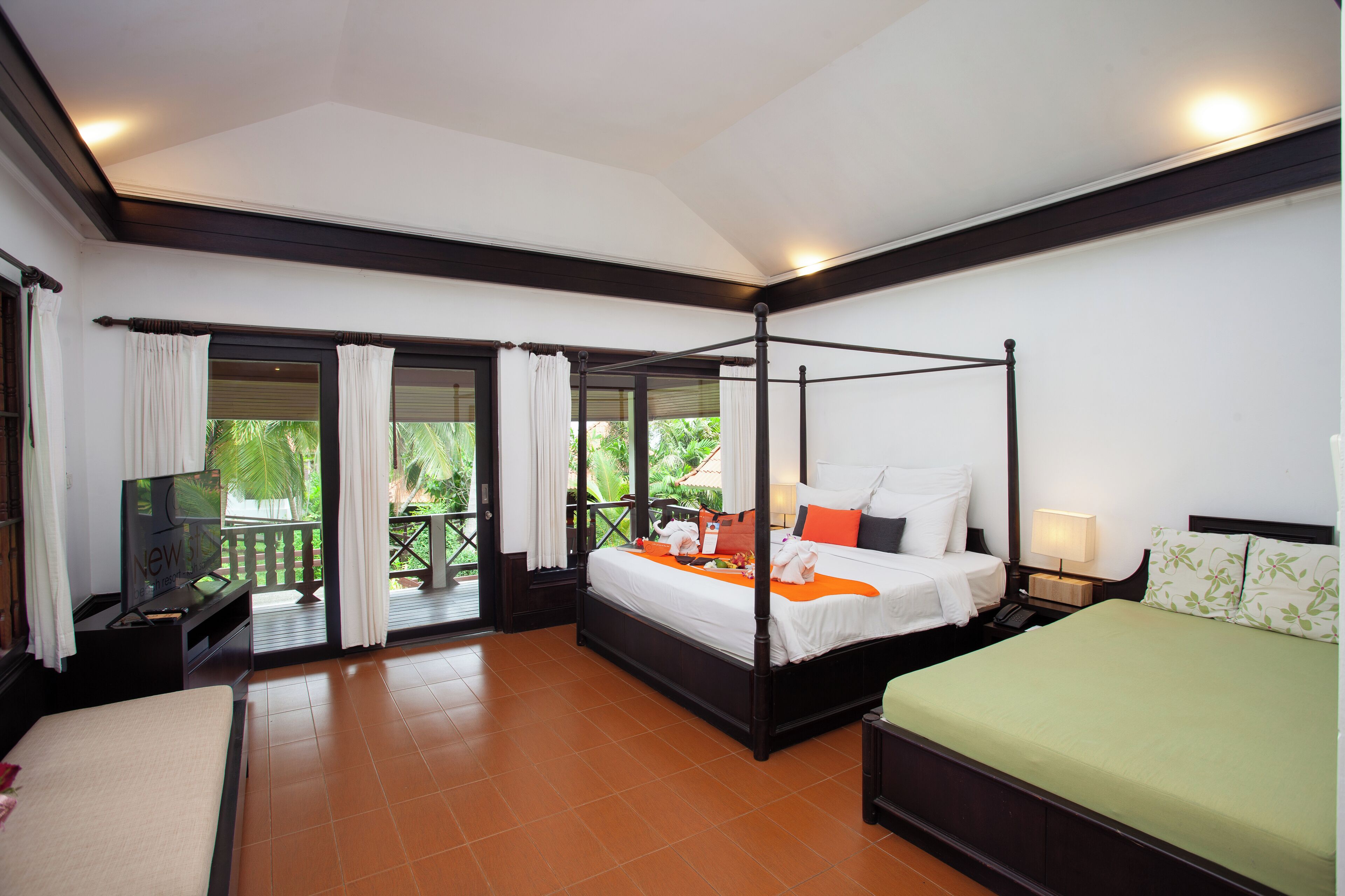 A four-poster bed with white bedding and orange accents, a television on a stand, and a balcony with curtains.