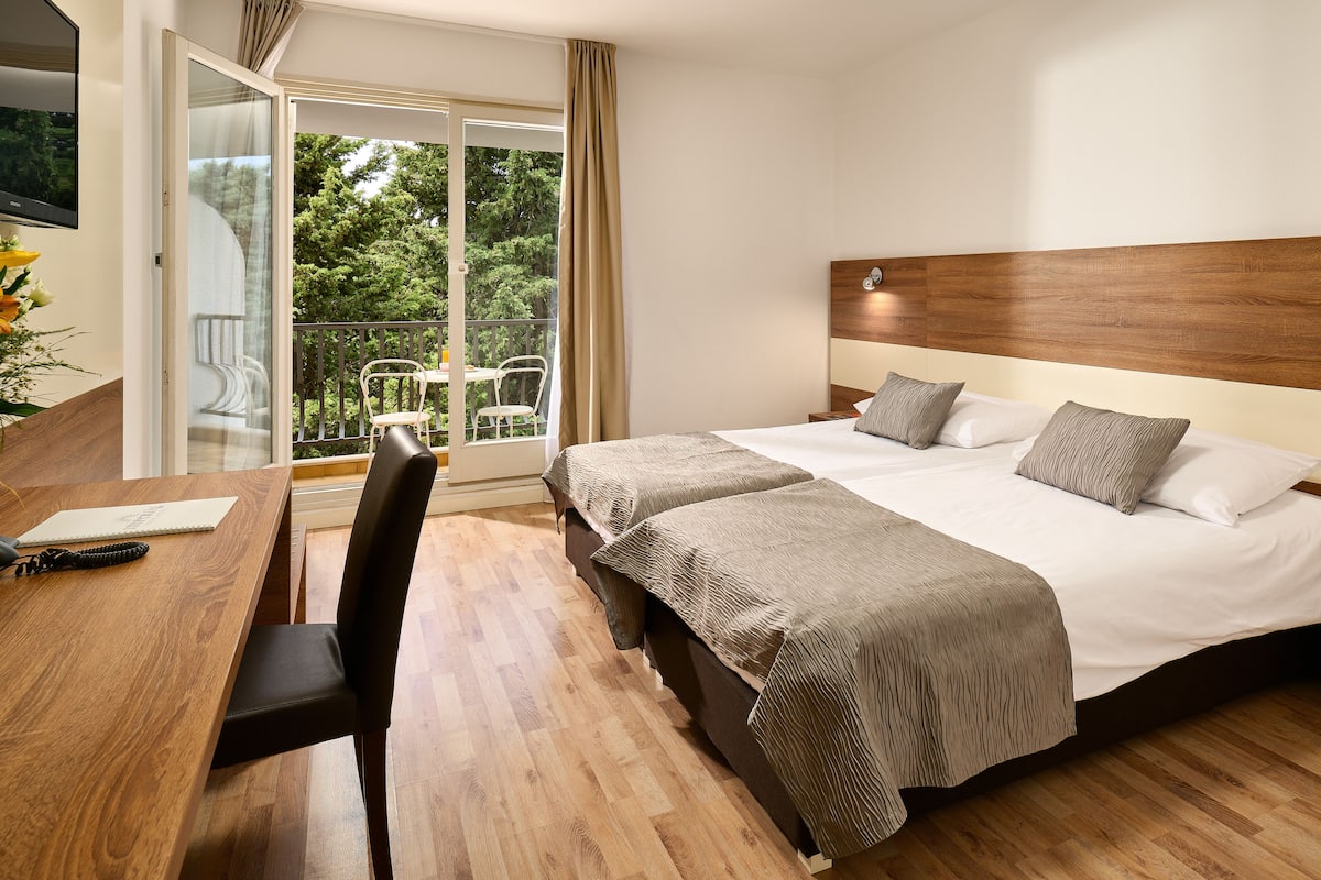 Habitación de hotel con piso de madera, una cama con dos almohadas, un escritorio con una silla y un balcón con mesa y sillas.