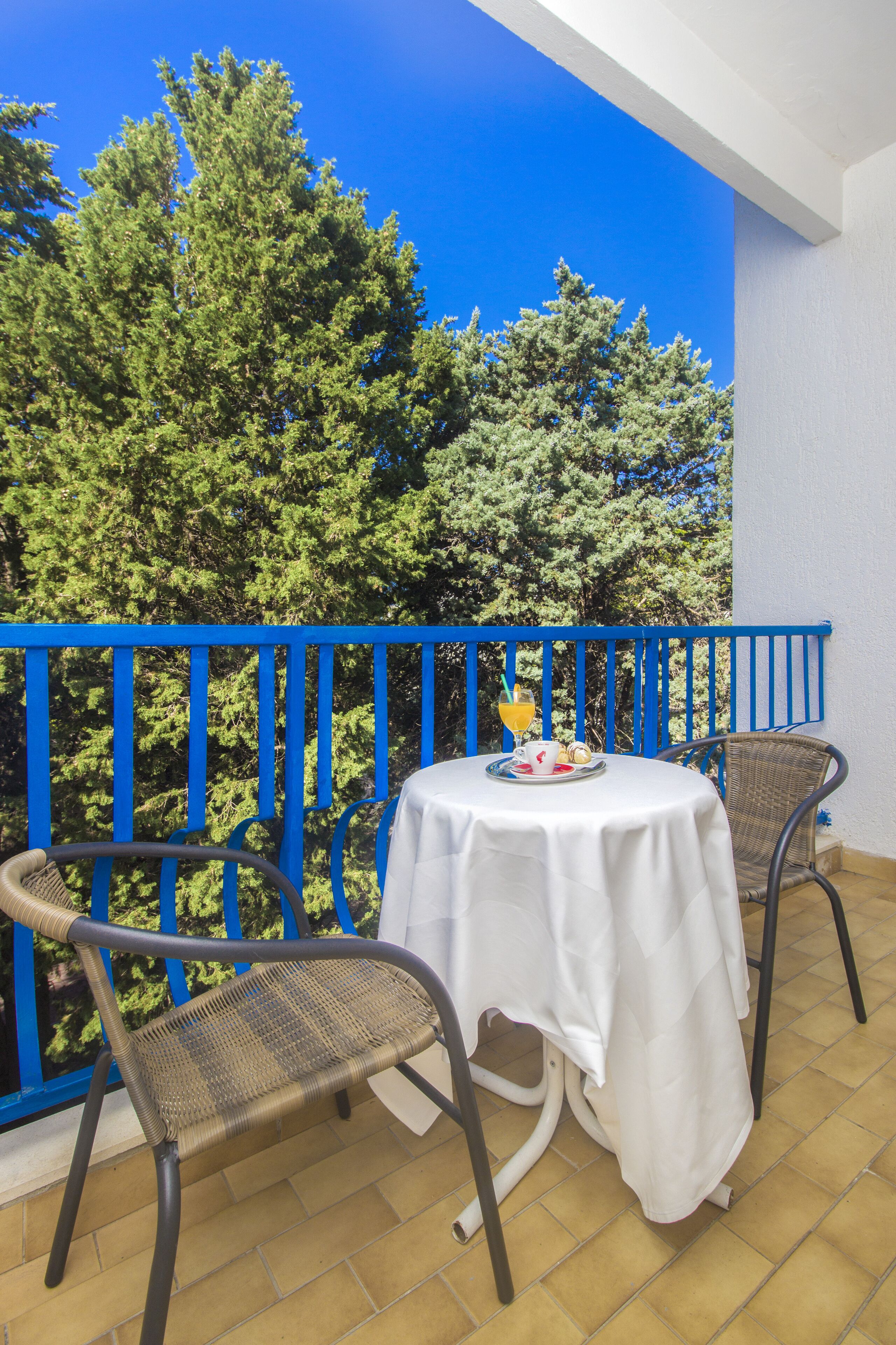 A balcony with a blue railing, a table with a white tablecloth, two wicker chairs, and a glass of orange juice.