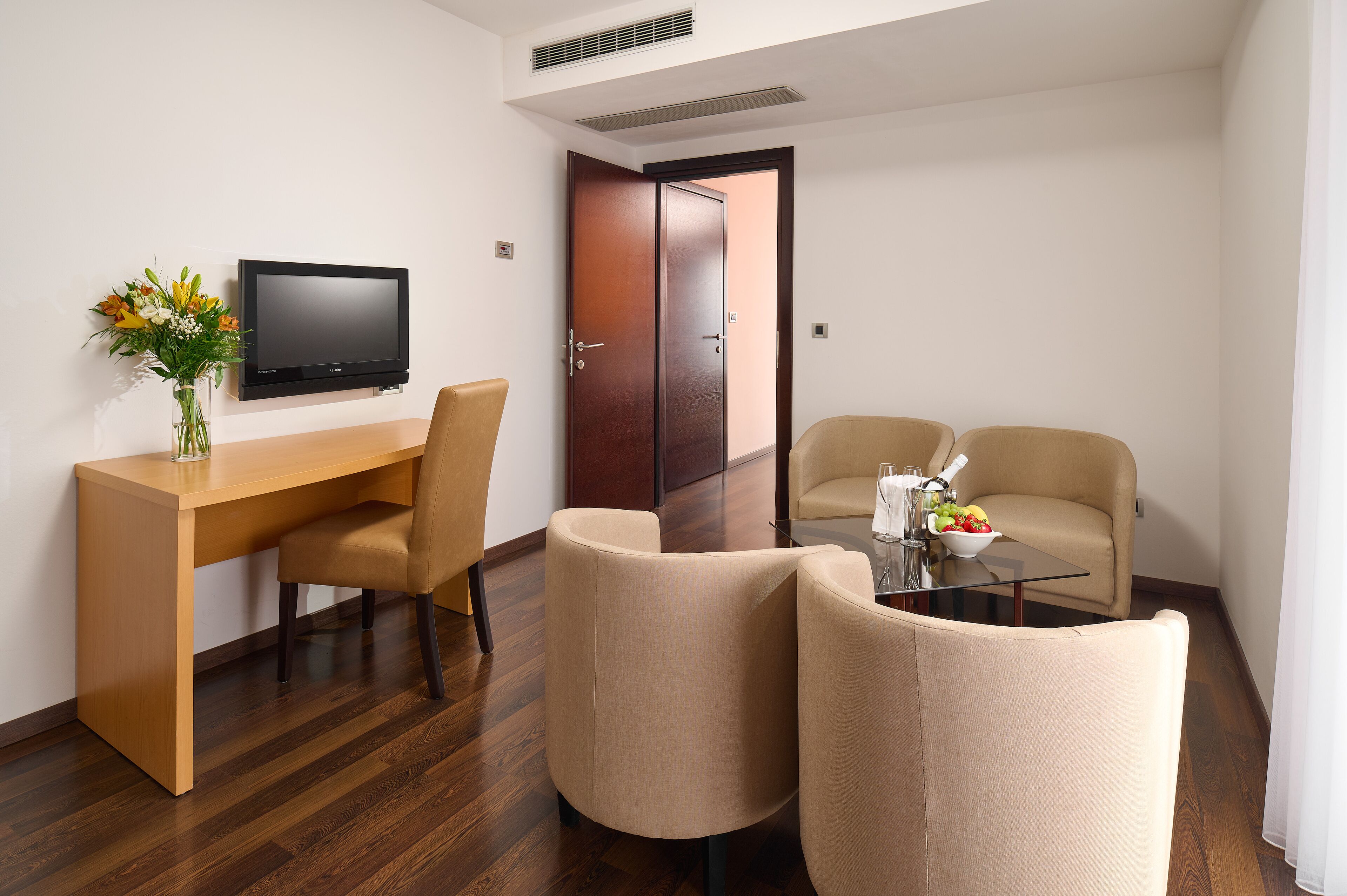 A hotel room with a wooden desk, a television, two beige armchairs, and a small table with a bowl of fruit.