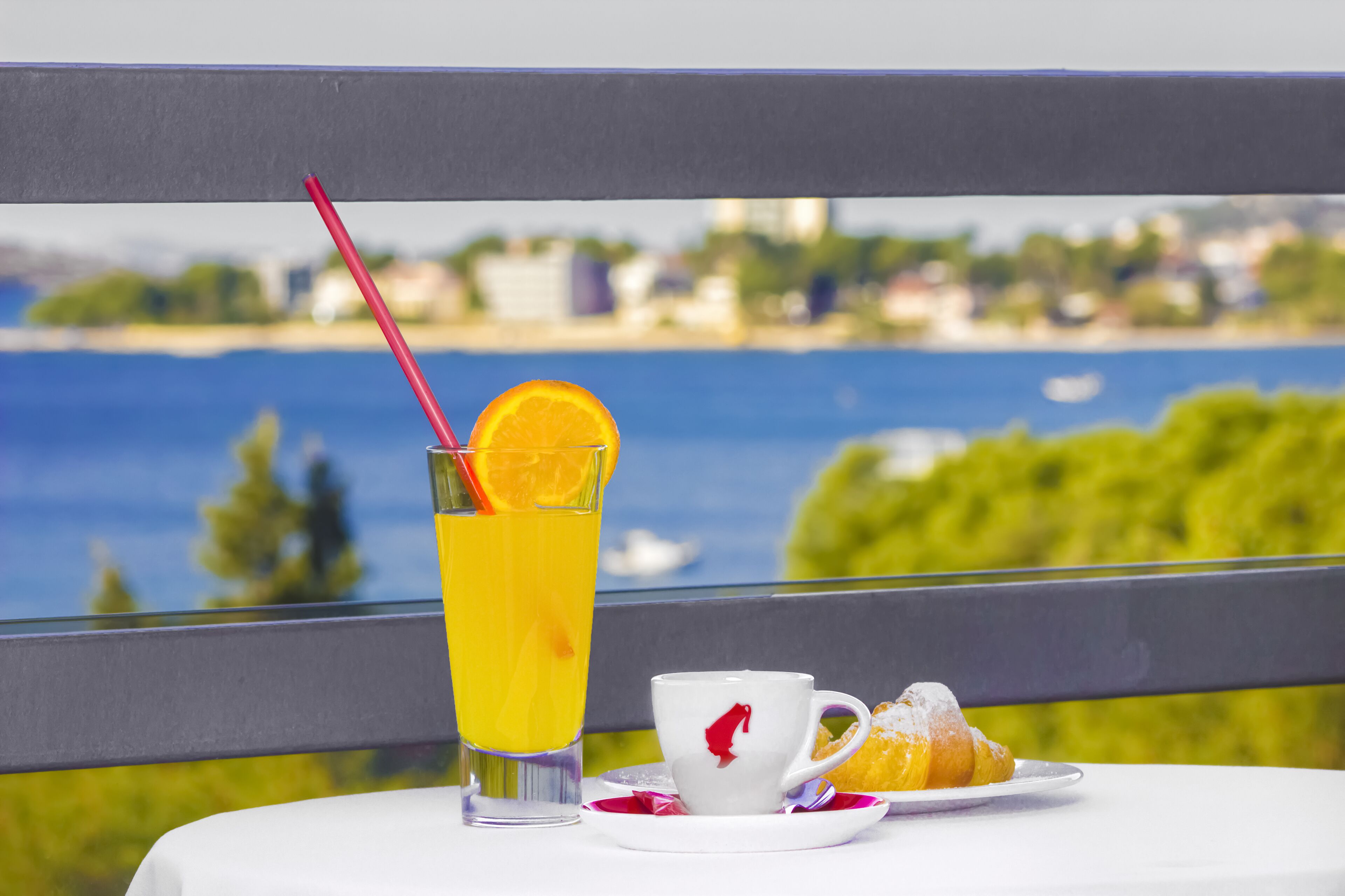 Ett bord med ett glas apelsinjuice, en kopp och ett bakverk, placerat med en vacker utsikt över en sjö eller ett annat vattendrag och grönskande omgivningar som bakgrund.