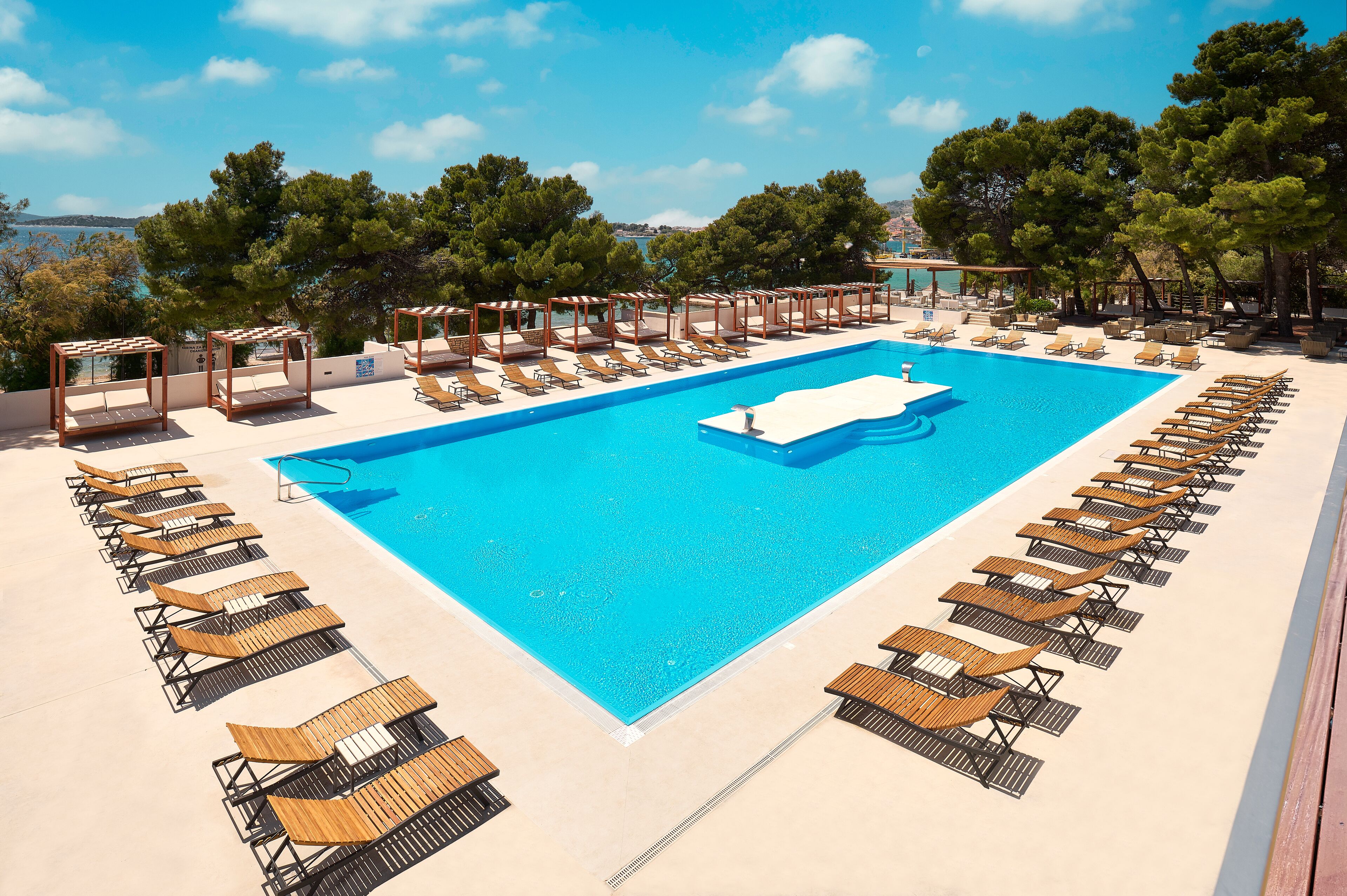 Une piscine dotée de chaises longues et d’une pergola, entourée d’arbres et offrant une vue sur la mer.