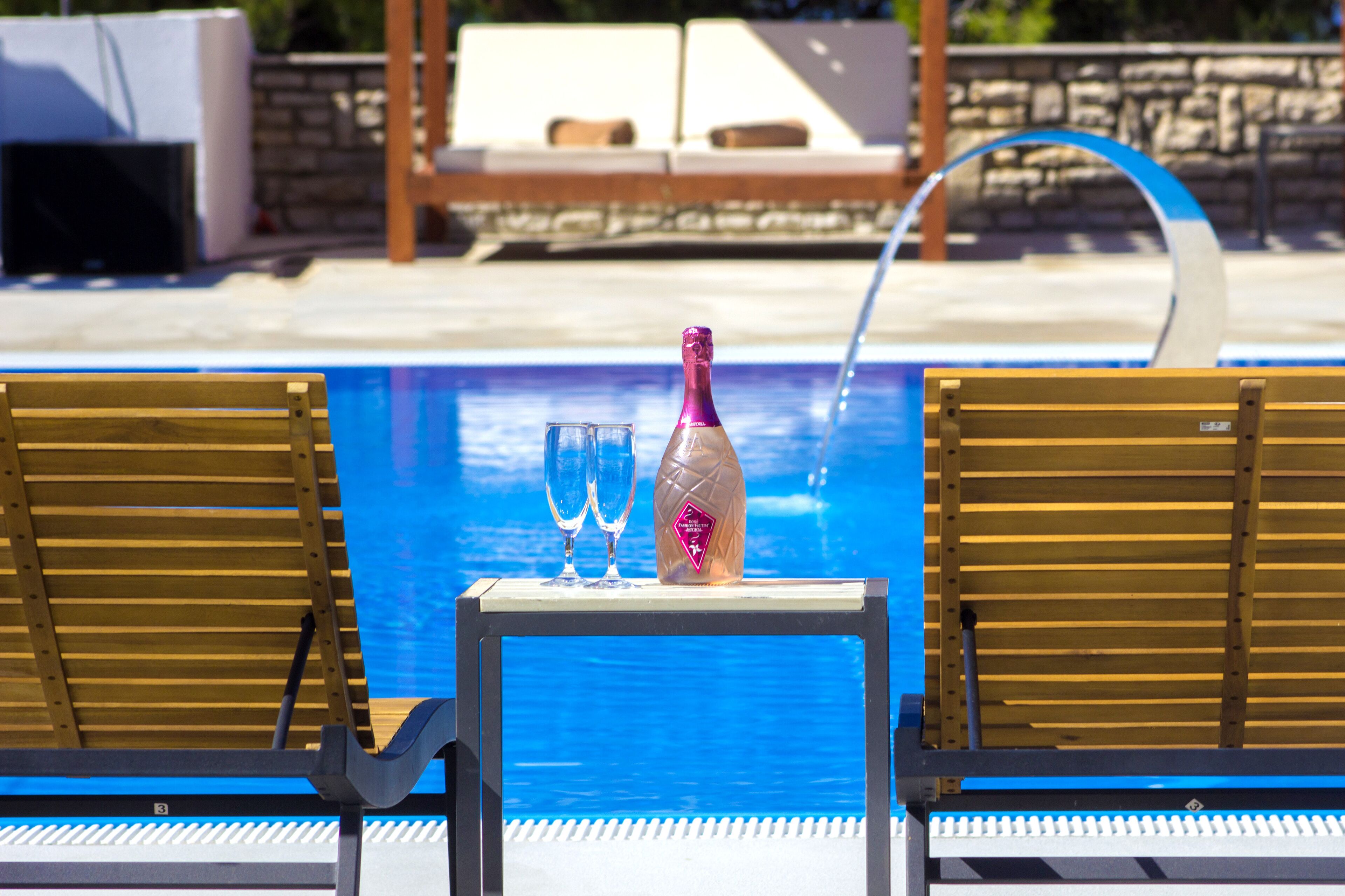 A poolside area with a table holding a bottle and two glasses, two wooden lounge chairs, and a stone wall in the background.