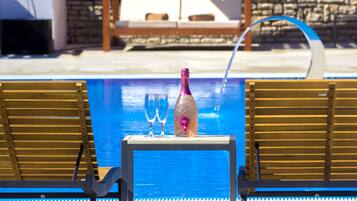 A poolside area with a table holding a bottle and two glasses, two wooden lounge chairs, and a stone wall in the background.
