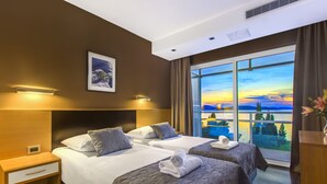A hotel room with two beds, a nightstand, a wall-mounted picture, and a large window with a scenic view. - Hotel Imperial Vodice (Vodice)