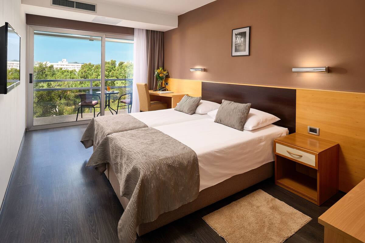 Habitación de hotel con una cama grande, un escritorio y una silla. Hay una puerta corrediza de vidrio que da a un balcón con vistas a árboles y edificios.