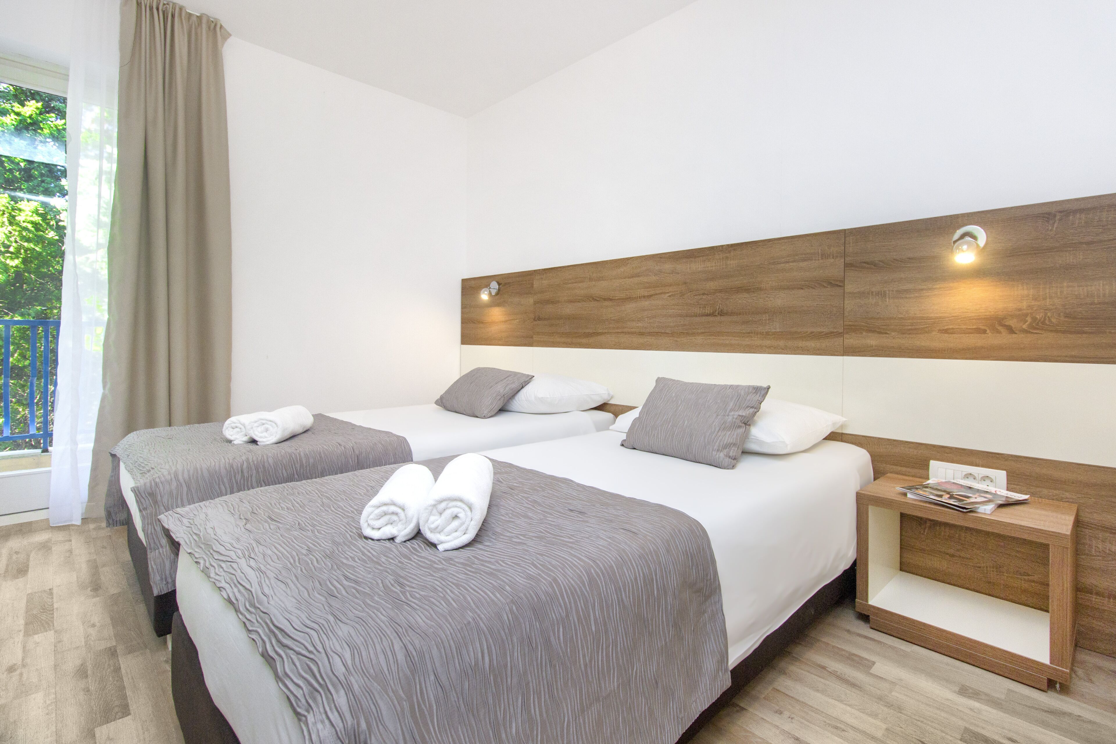 Hotellihuone, jossa on kaksi sänkyä, puinen pääty ja yöpöytä, jonka päällä on lehti.