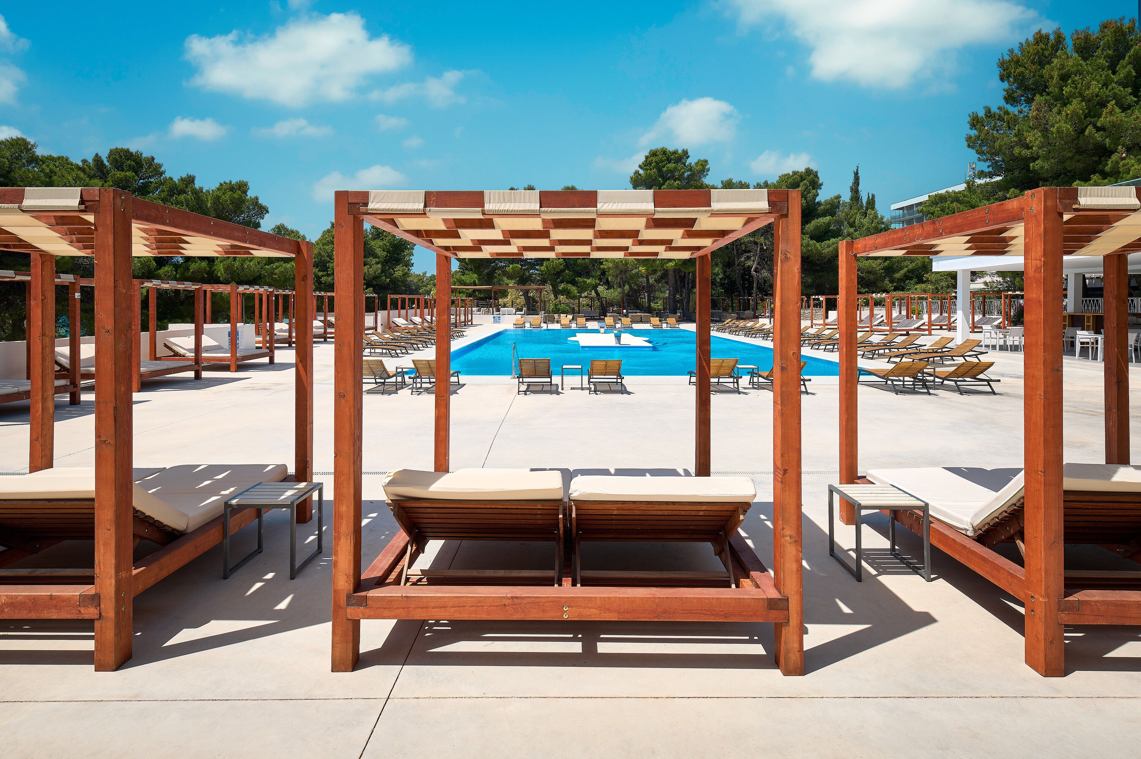 A poolside area with wooden loungers and umbrellas, a clear blue pool, and a view of trees and buildings in the background.