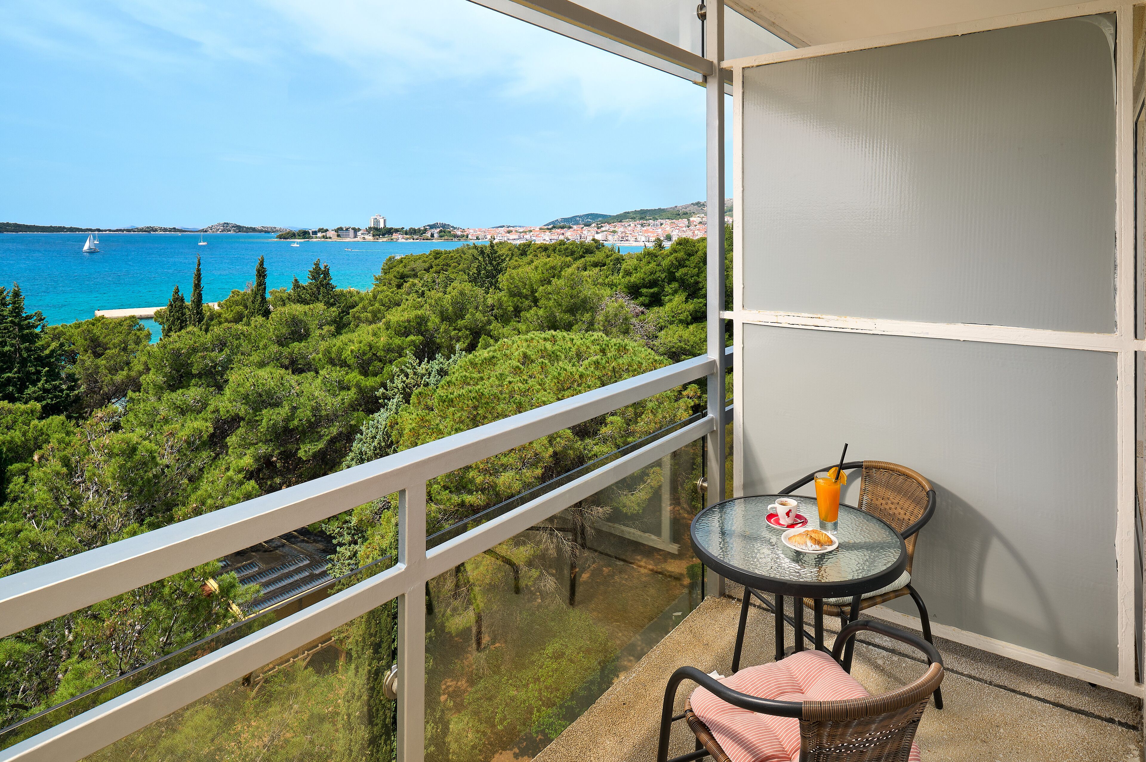 Un balcon avec une table et des chaises, donnant sur une zone côtière bordée d’arbres et offrant une vue sur la mer.