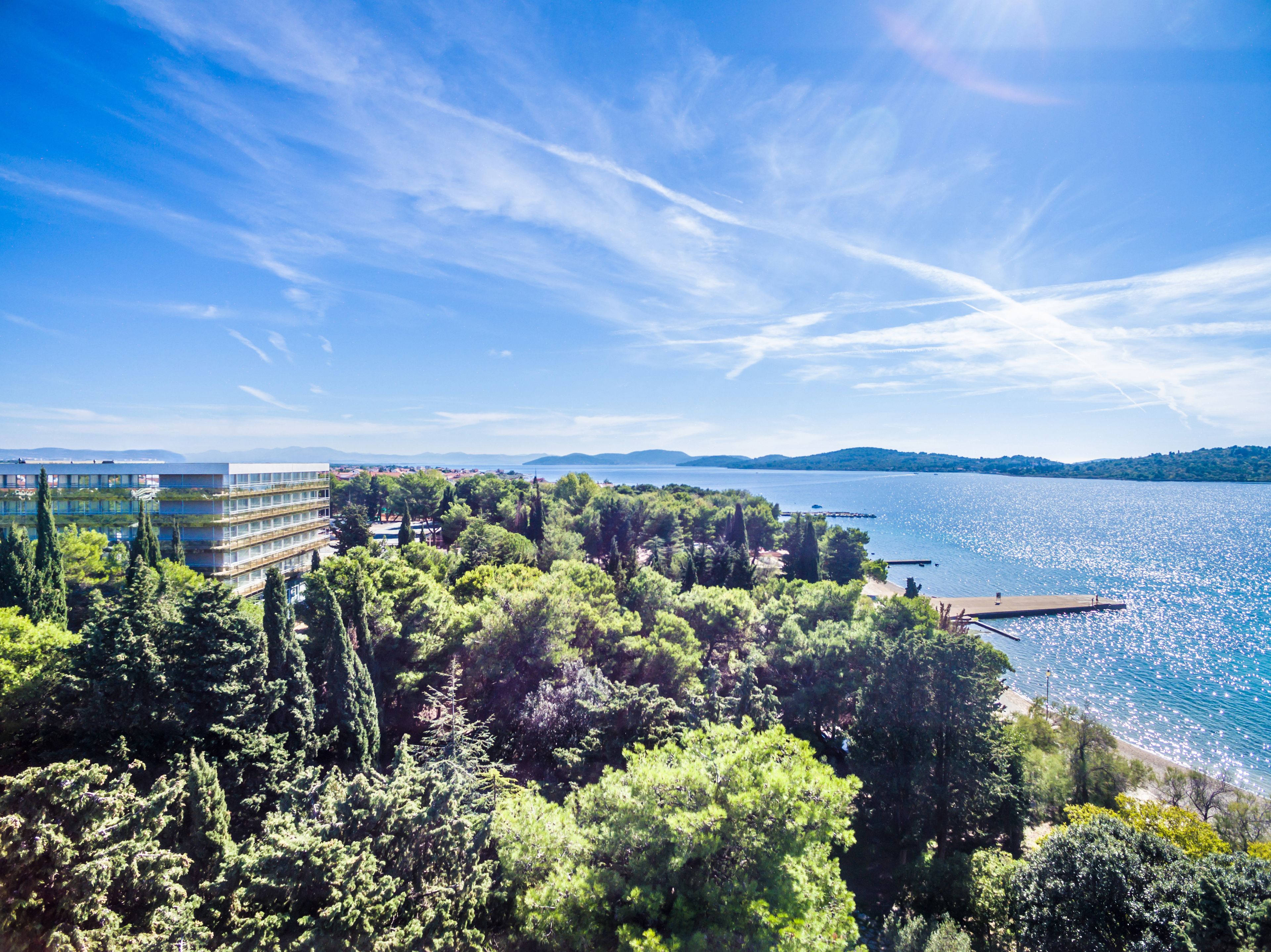 A coastal landscape with a modern building, dense greenery, and a clear blue sky.