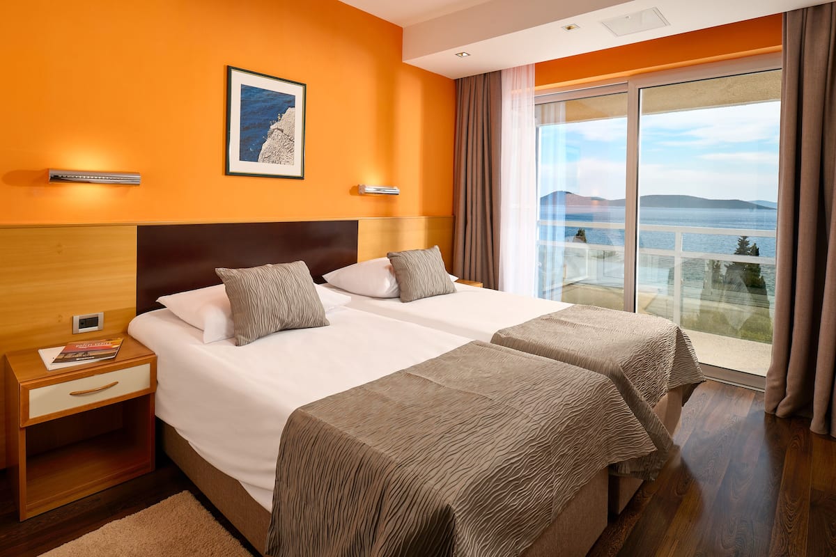 Habitación de hotel con dos camas, una mesita de noche, un cuadro enmarcado y un amplio ventanal con vistas al mar.