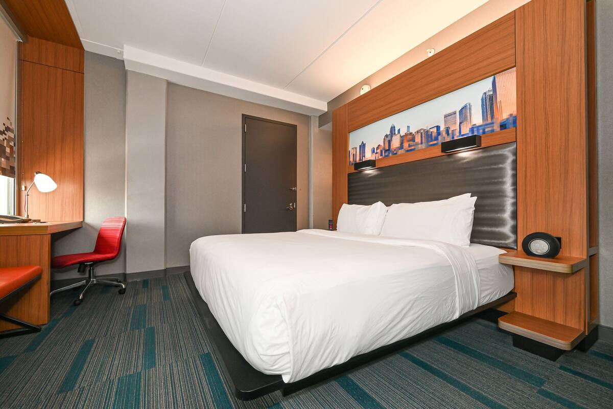 A hotel room with a large bed, a desk with a chair, a cityscape painting on the wall, and a door.