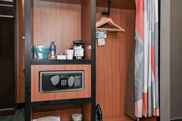 Habitación de hotel con pared de paneles de madera, minibar con botella de agua, cafetera, artículos de aseo, panel de control y perchero.