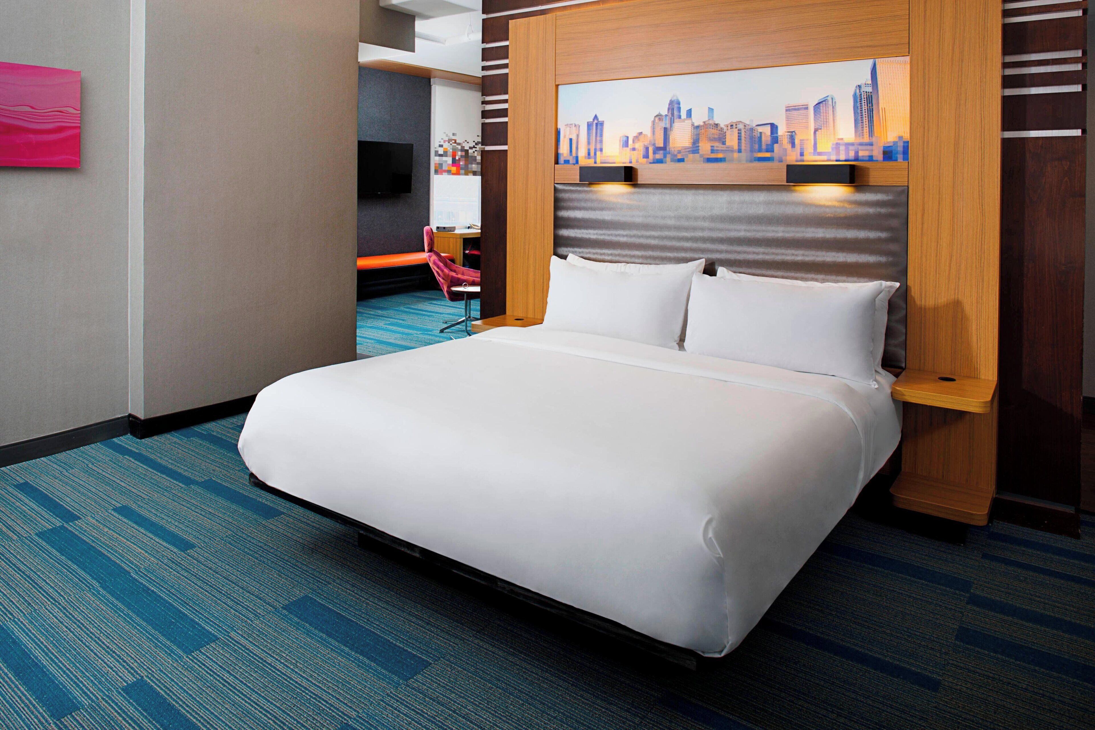 A hotel room with a large bed, a cityscape painting on the headboard, a desk with a chair, and a television.