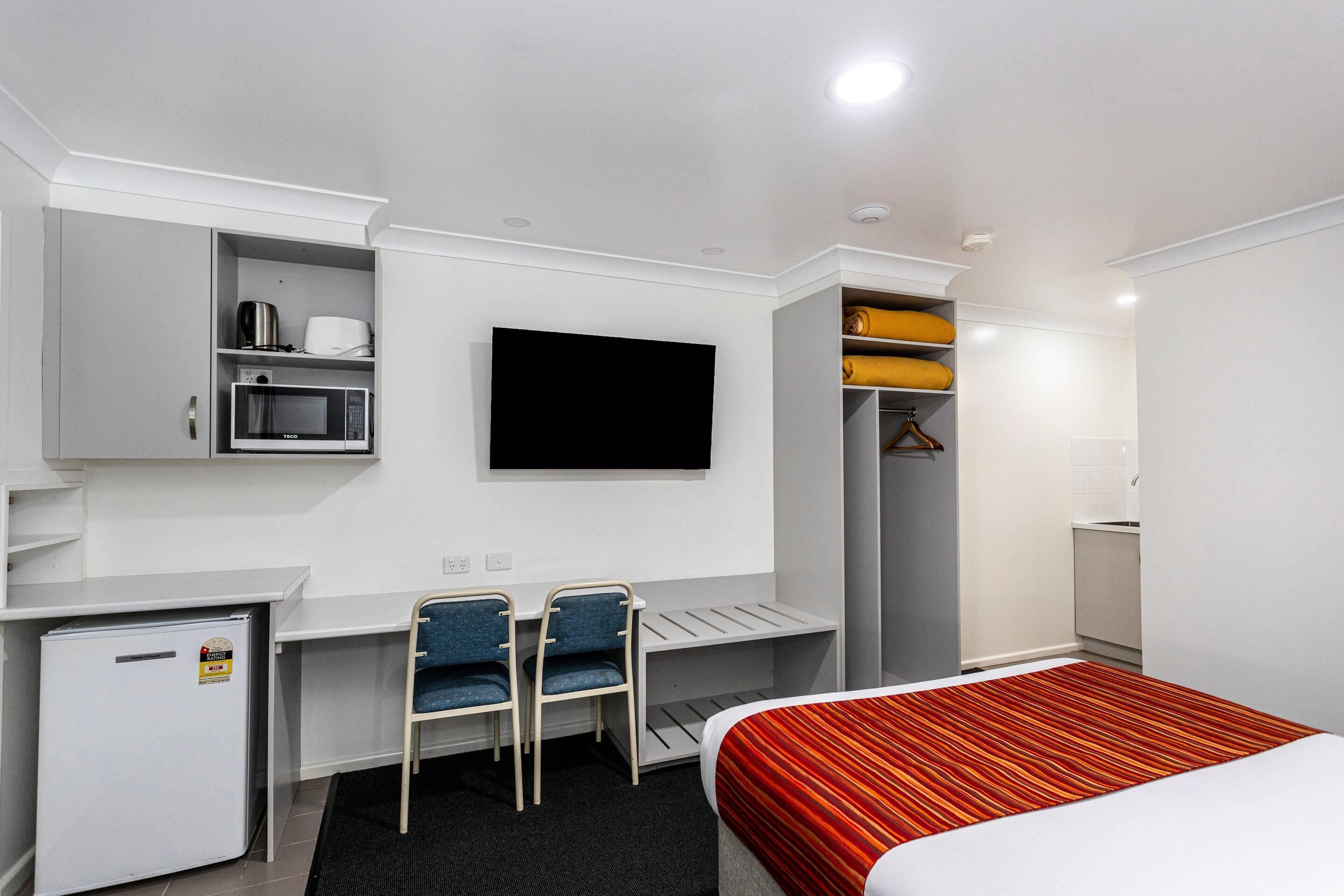 Habitación de hotel con cama, escritorio con silla, televisor, refrigerador pequeño y armario.