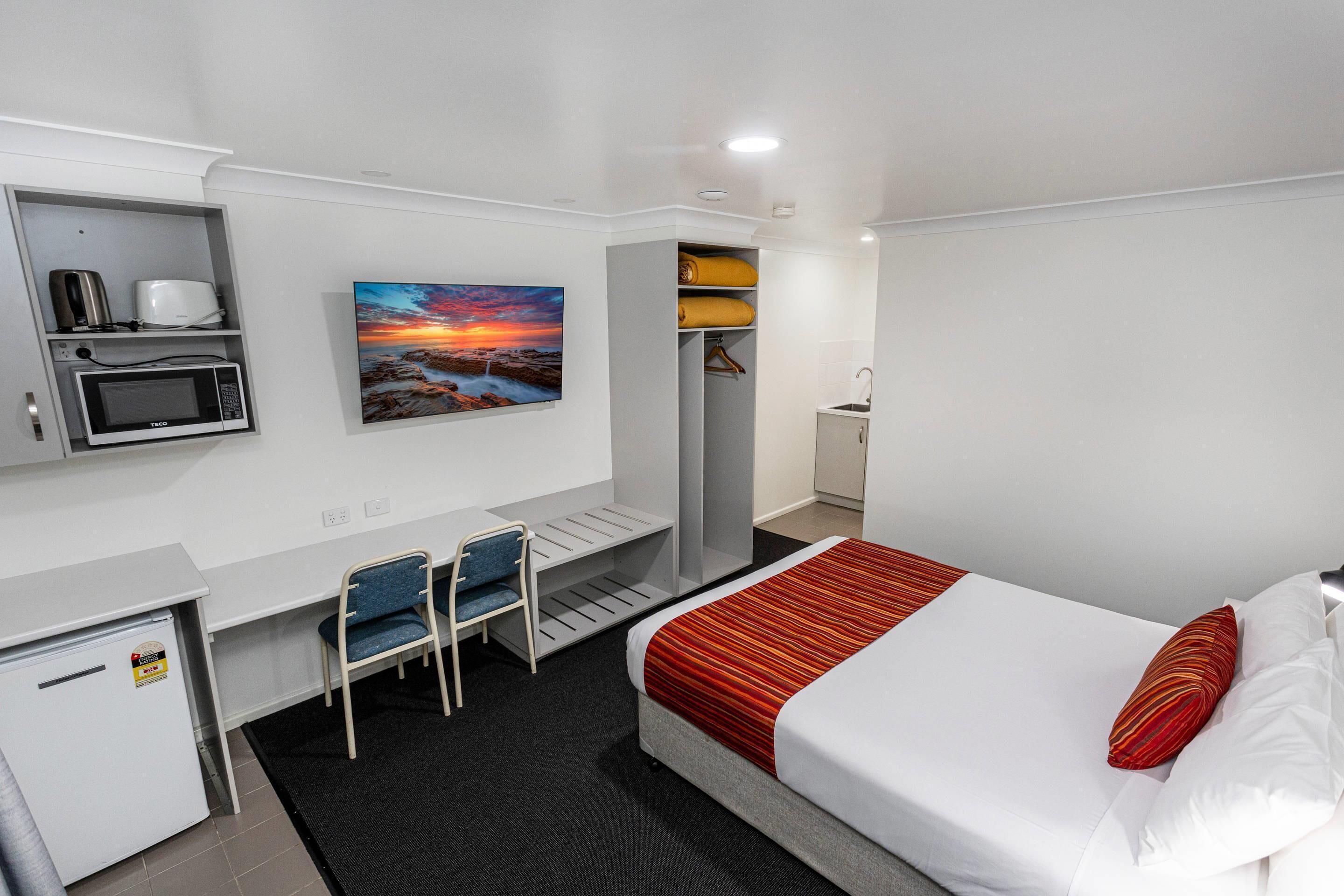 A hotel room with a bed, a desk with a chair, a microwave, a small fridge, a painting, and a wardrobe.