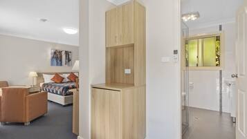Habitación de hotel con cama, armario de madera, zona de estar con una silla y baño con ducha de vidrio.