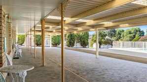 Un espace extérieur abrité, doté de tables et de chaises, d’un sol en béton et offrant une vue sur des arbres et un bâtiment en arrière-plan.