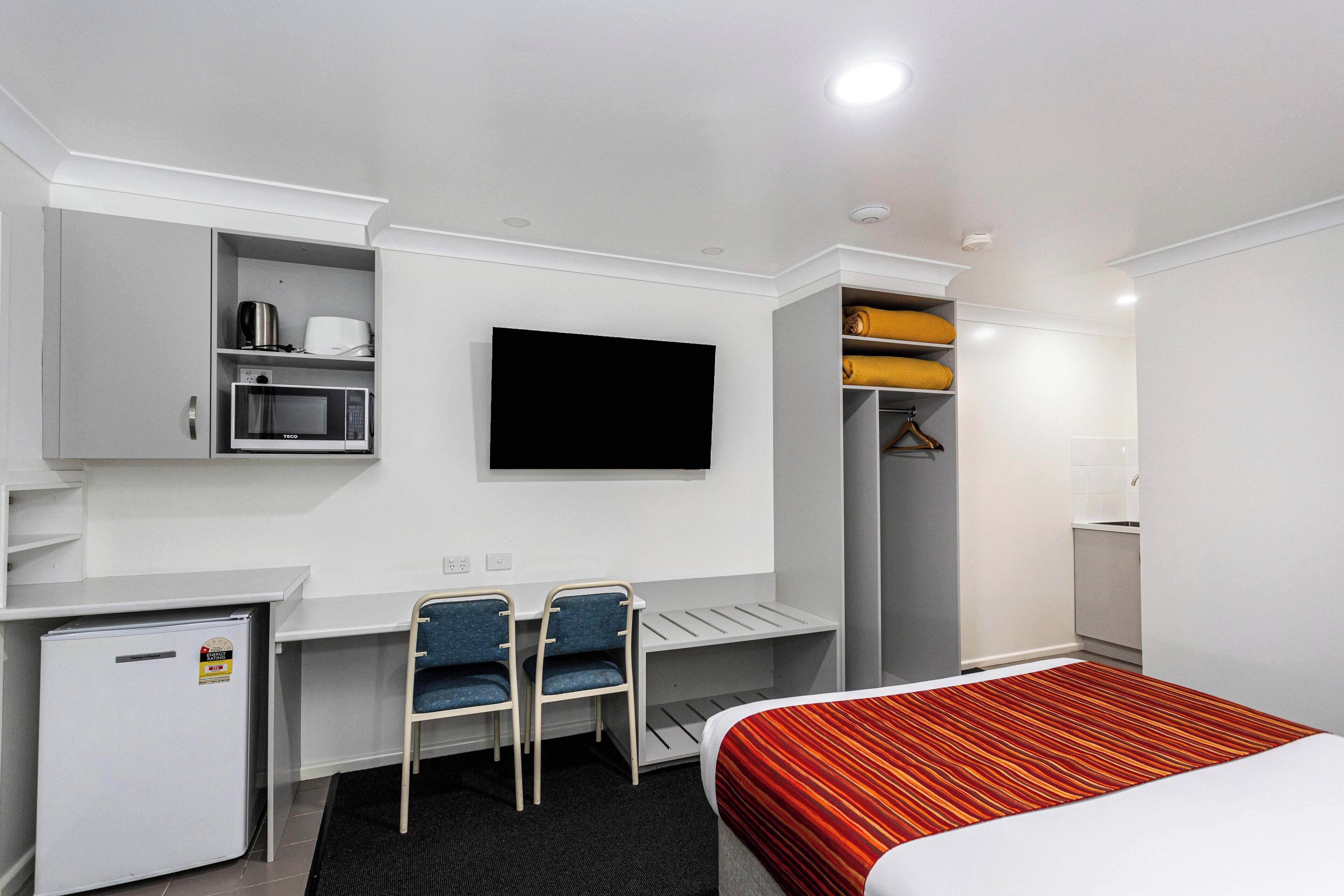 A hotel room with a bed, desk, chairs, a TV, a mini-fridge, and a wardrobe.