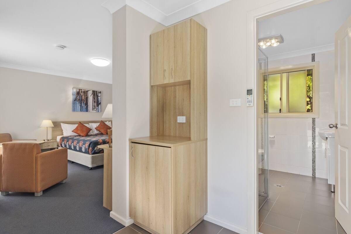 A hotel room with a bed, a wooden wardrobe, a bathroom with a glass shower, and a seating area with a chair.