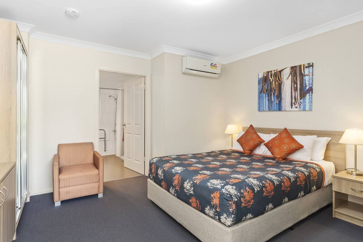 A hotel room with a bed, a chair, a nightstand, a wall-mounted air conditioner, and a door leading to another room.