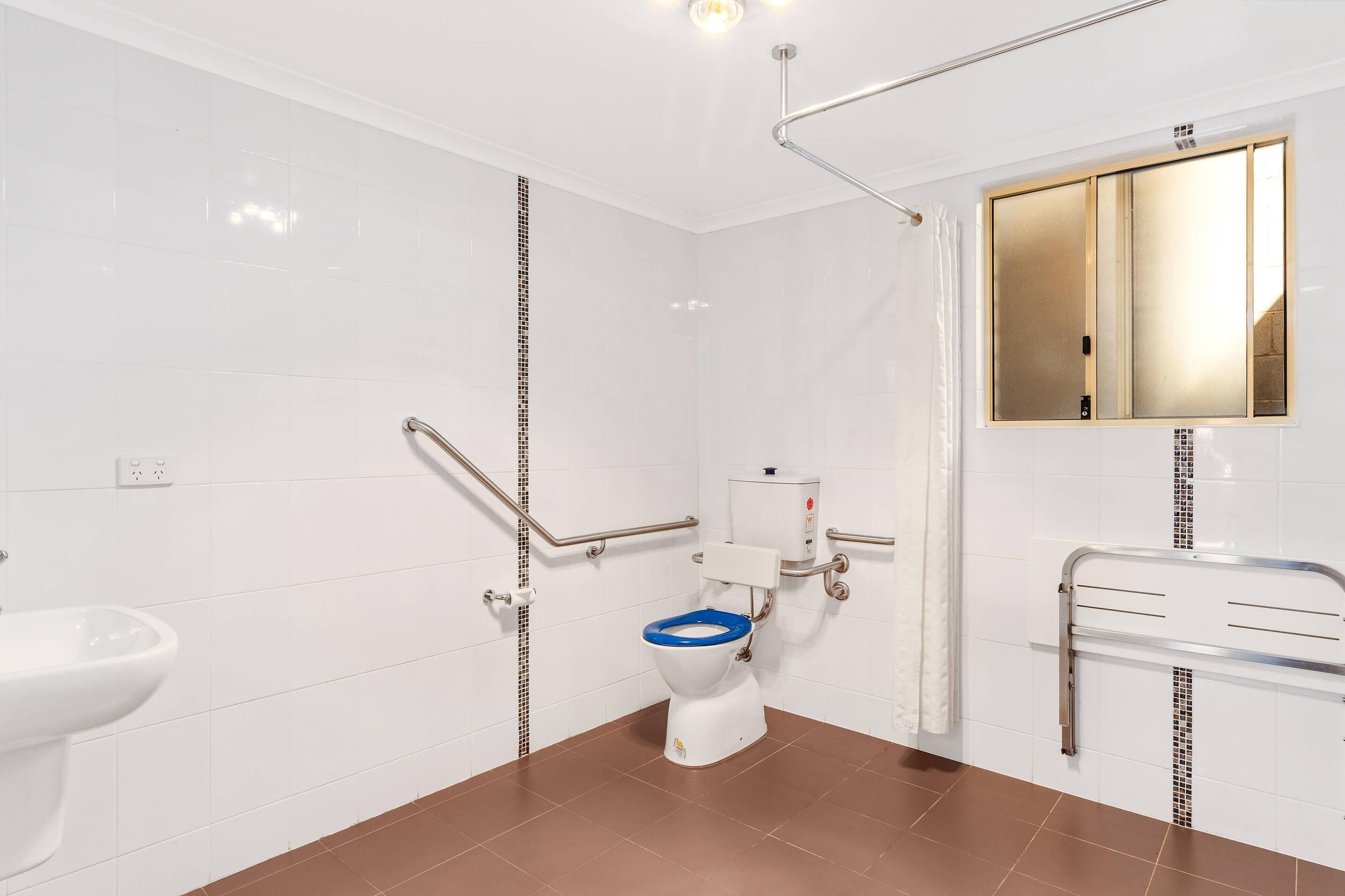 A bathroom with white tiled walls, a brown floor, a white toilet with a blue seat, a white sink, and a mirror above the sink.