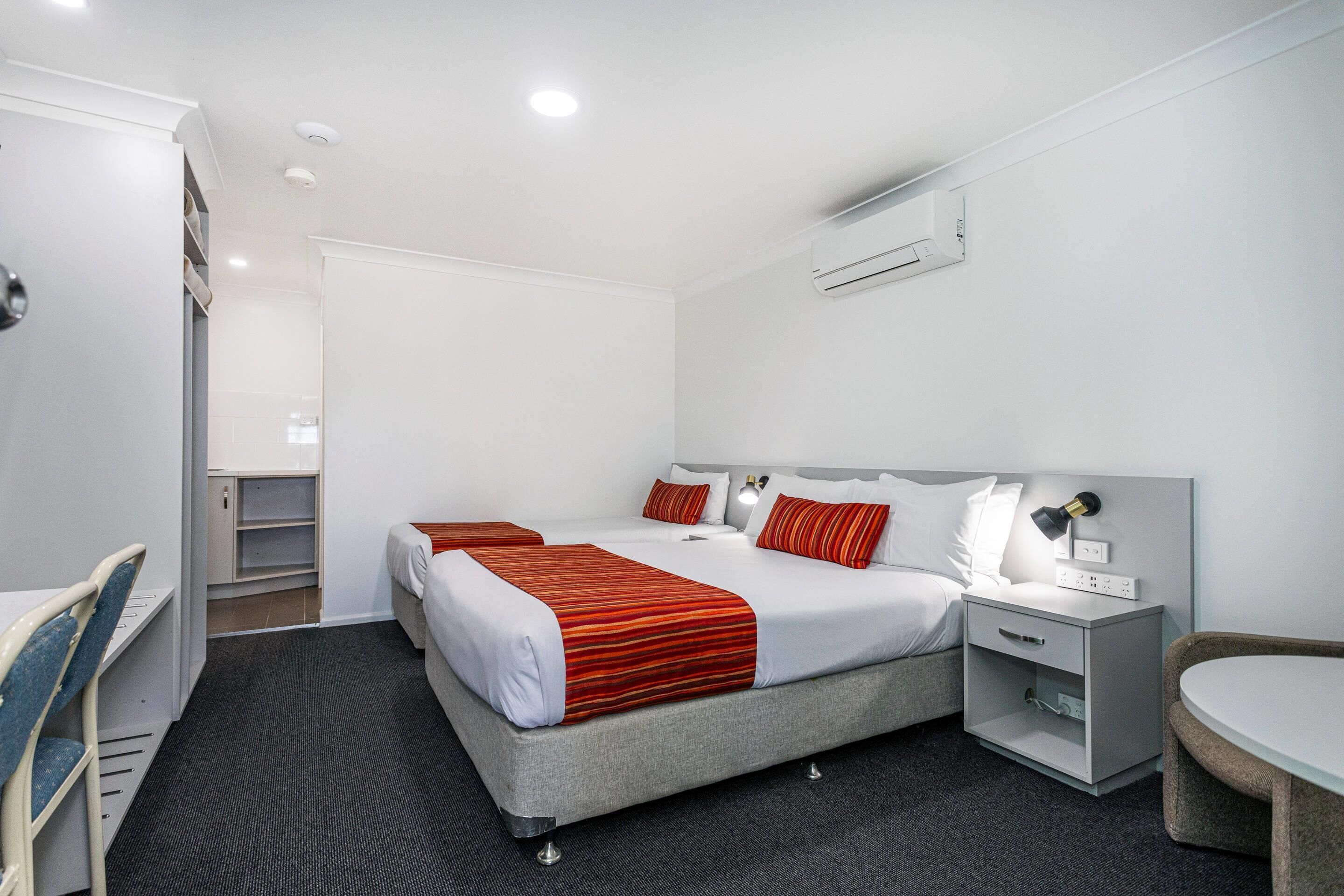 A modern hotel room with two beds, a desk, a chair, a bedside table with a lamp, and an air conditioning unit.