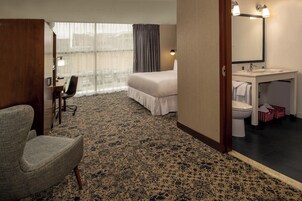 A hotel room with a bed, a desk with a chair, a bathroom with a sink and toilet, and a large window with curtains.