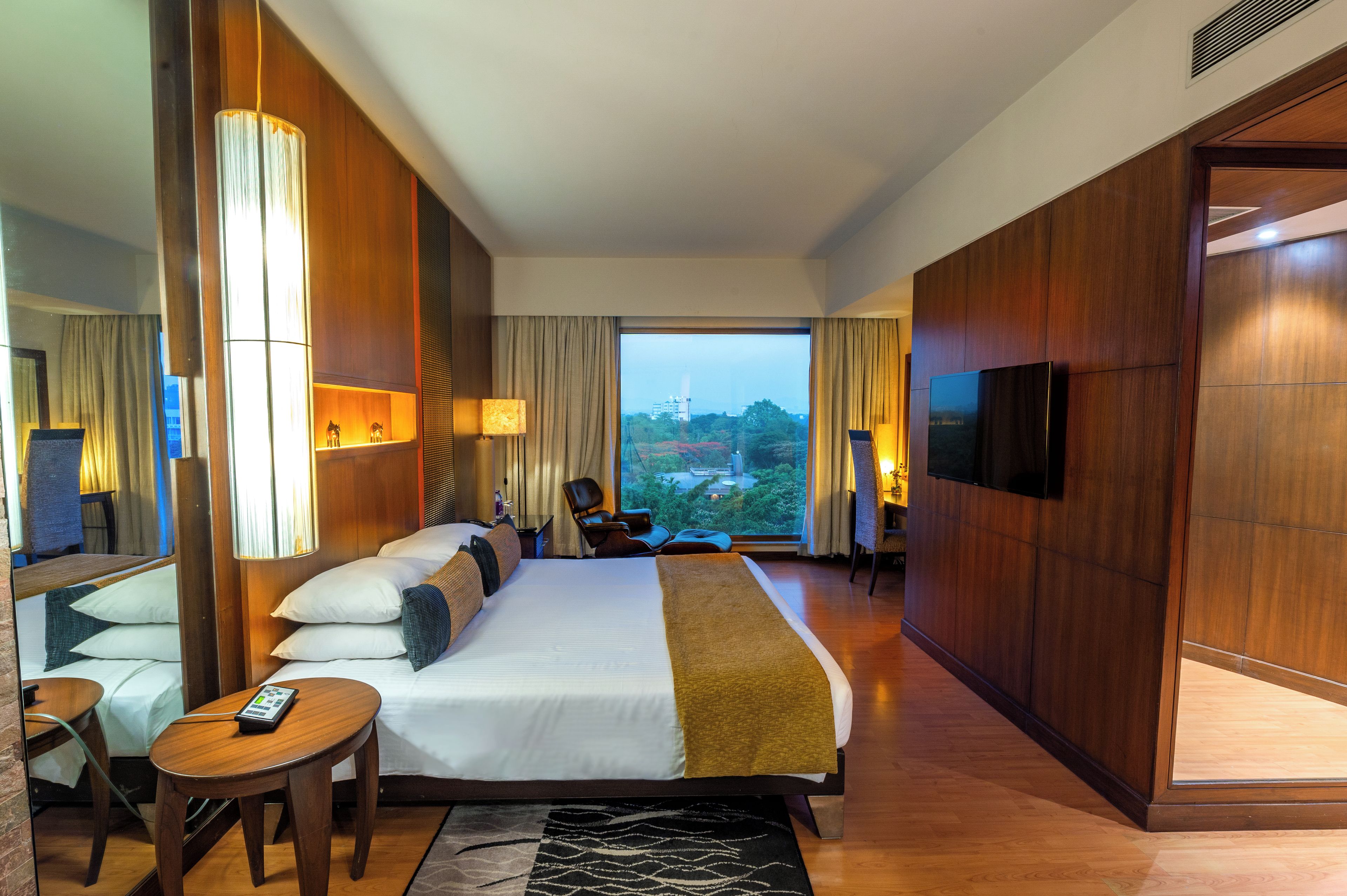 A hotel room with a large bed, a desk, a chair, a TV, and a view of a cityscape.