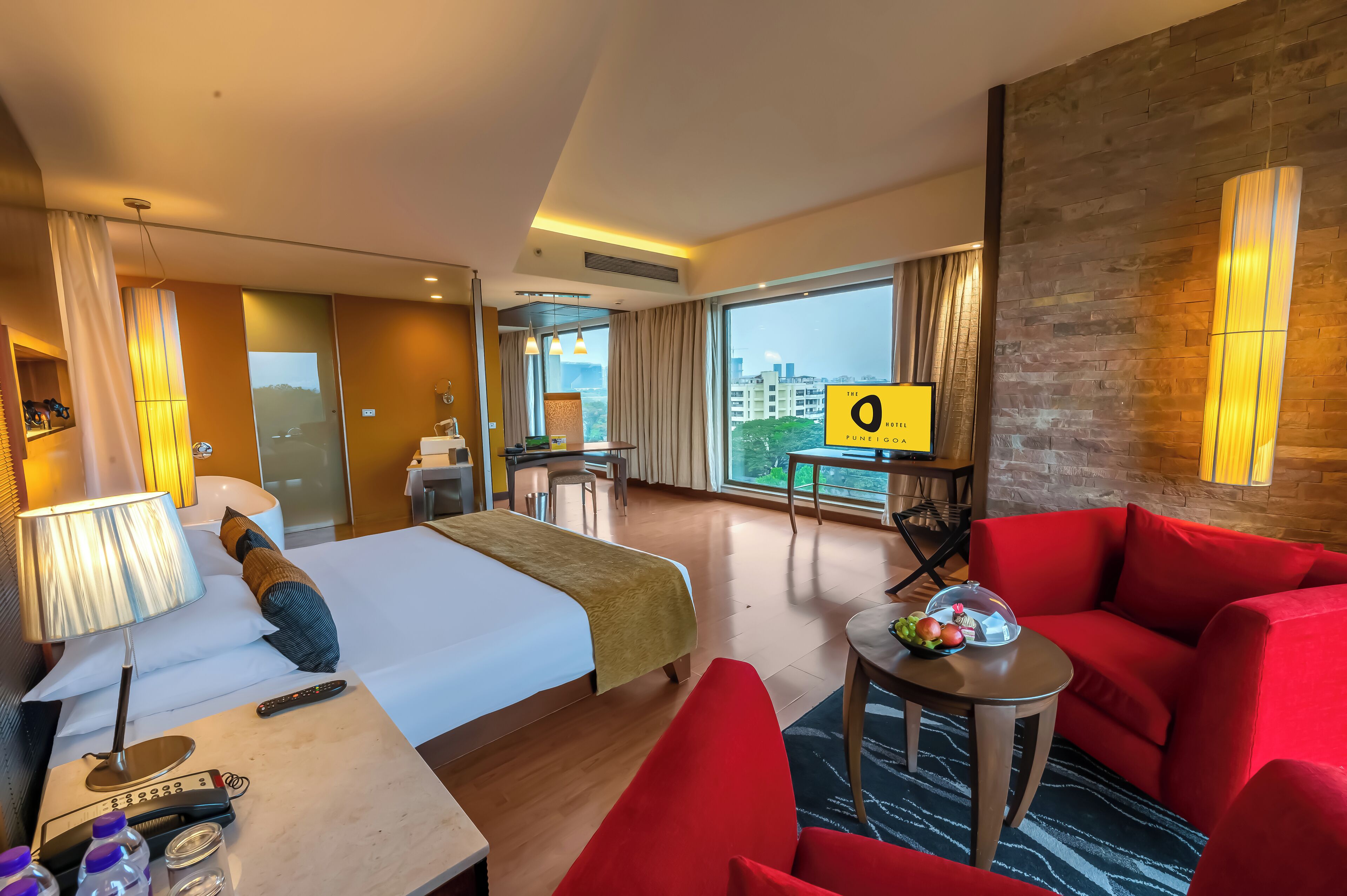 Una camera d'albergo con un letto, una scrivania, un divano rosso, un tavolino con una ciotola di frutta e vista sulla città attraverso ampie finestre.